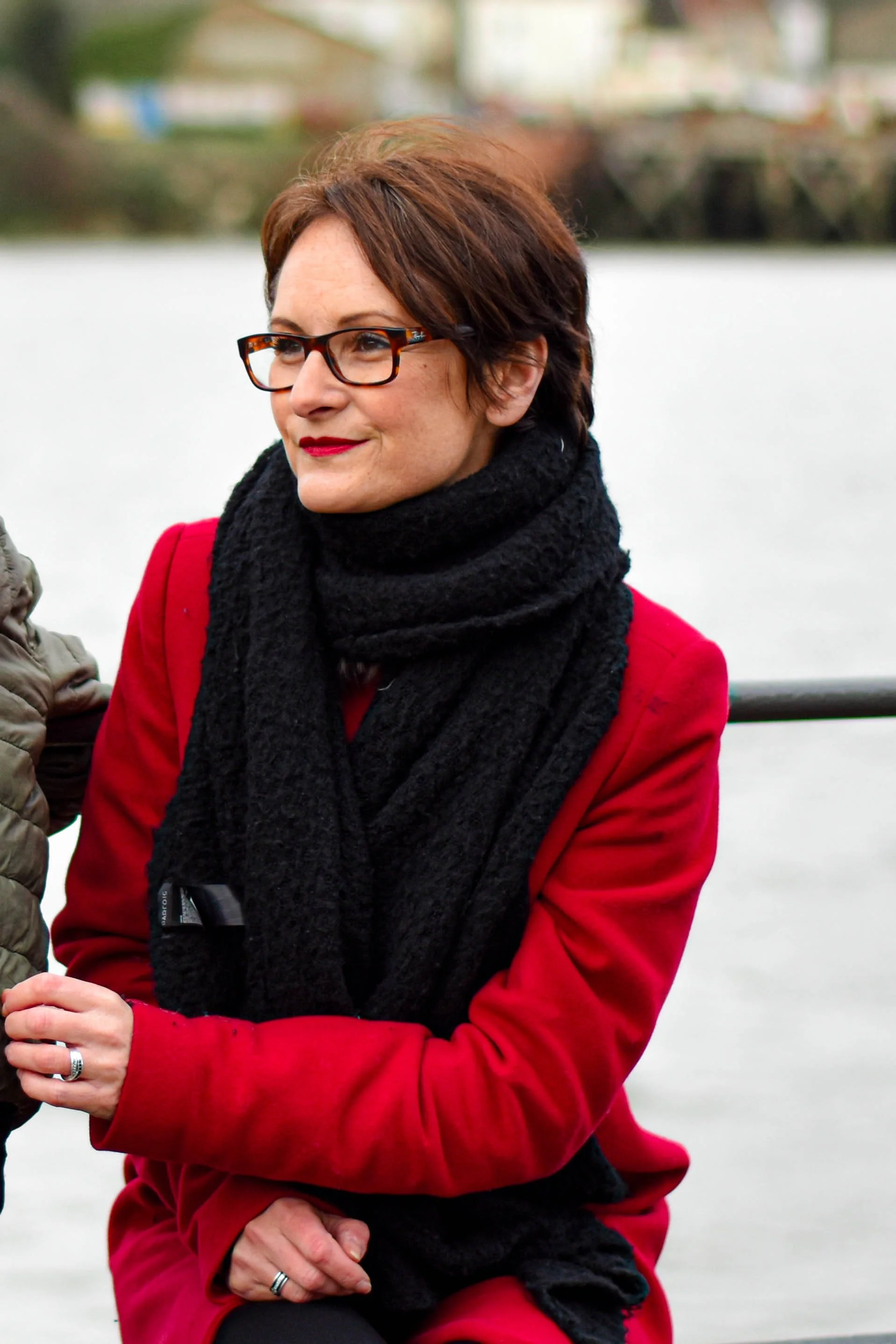 Une femme portant des lunettes à monture écaille et un manteau rouge, avec une écharpe noire, assise près d'un plan d'eau.