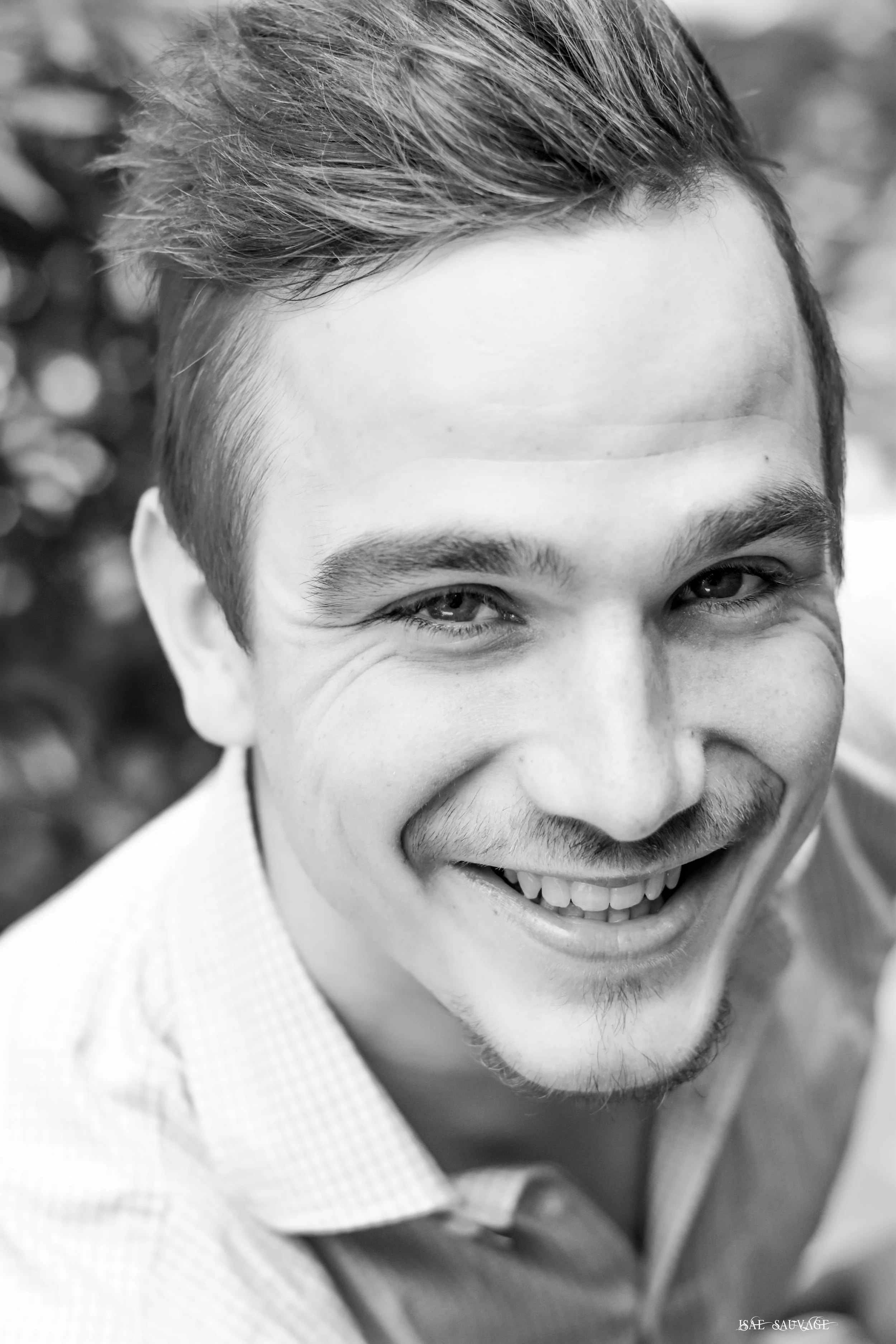 Un homme souriant avec des cheveux courts et une barbe, portant une chemise à carreaux. Photo en noir et blanc, partie supérieure du visage visible.