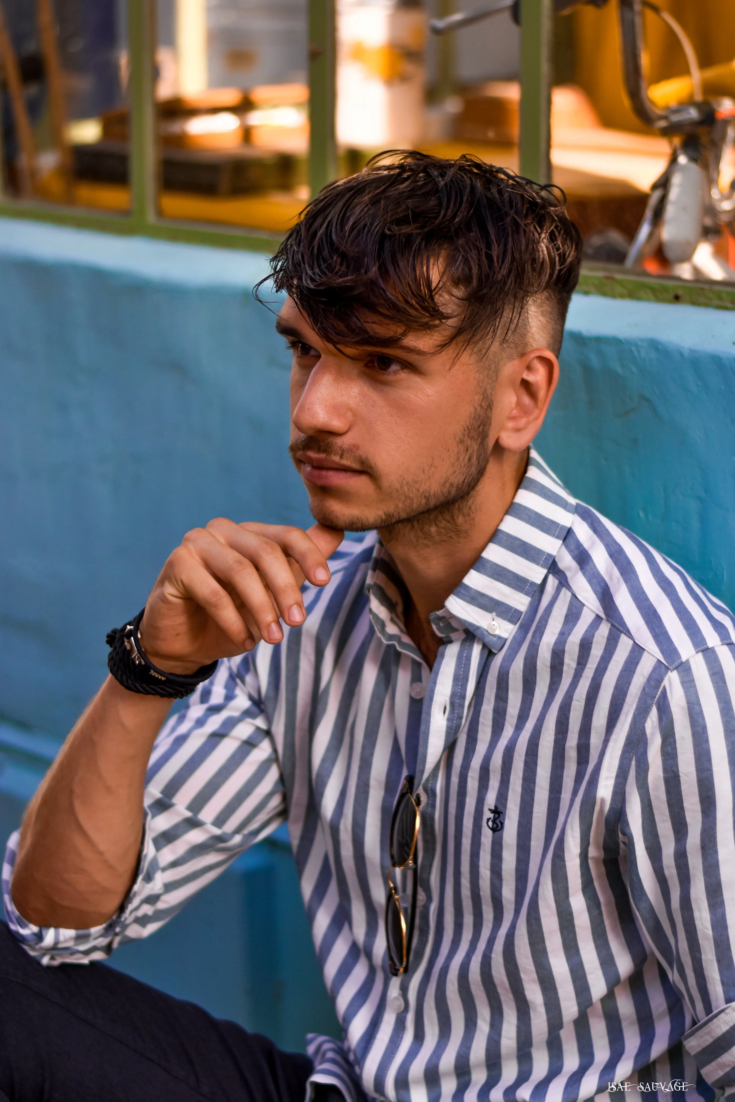 Un jeune homme avec les cheveux bruns bouclés, portant une chemise à rayures bleues et blanches, regarde pensivement avec la main sous le menton dans un décor urbain.