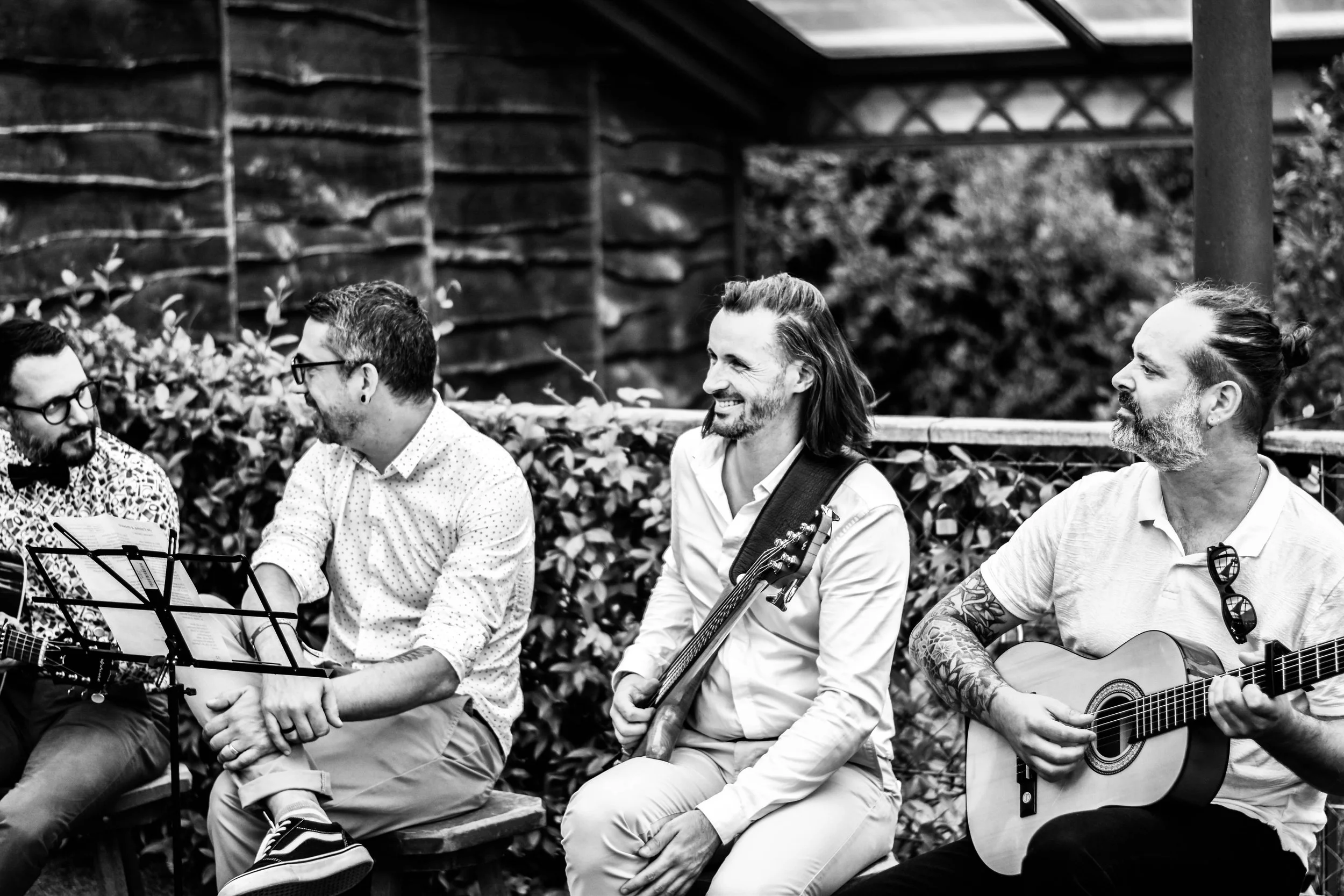 Groupe de quatre hommes assis sur un banc, jouant de la musique avec des guitares, dans un cadre extérieur avec des plantes et une structure en bois en arrière-plan.