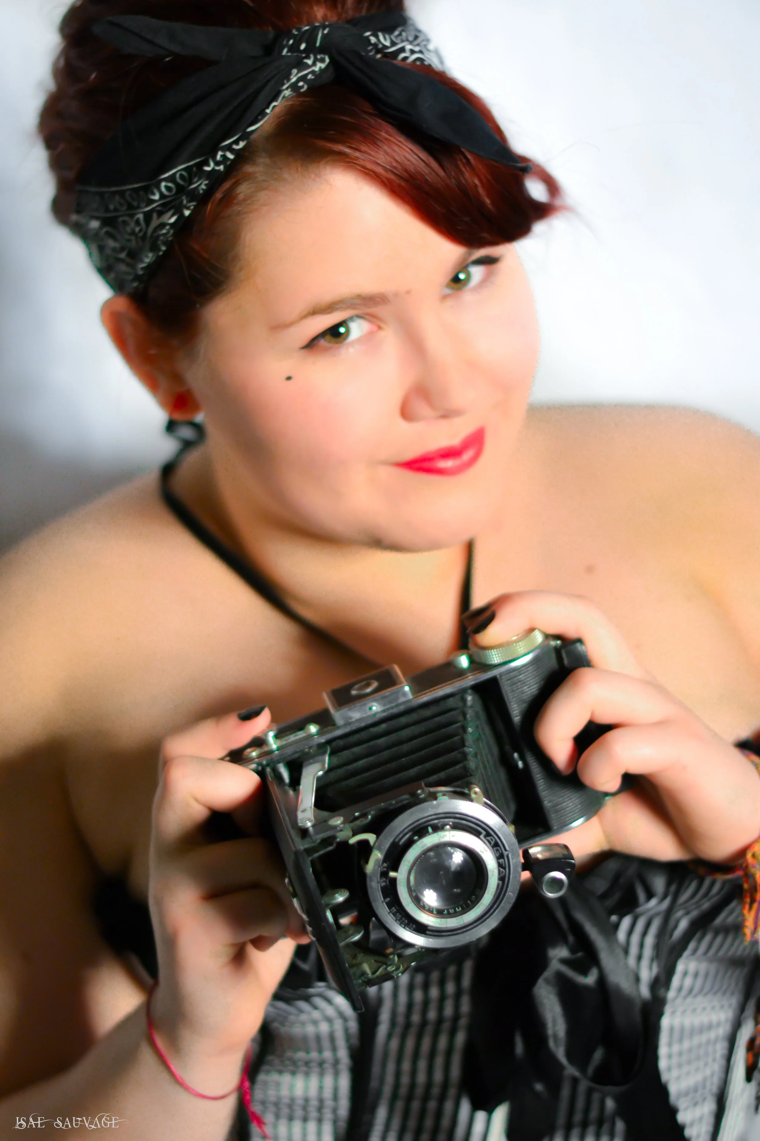 Une jeune femme avec des cheveux courts et rouges, portant un bandana noir et blanc, tient un appareil photo vintage en main, regardant vers l'objectif, avec un fond blanc.