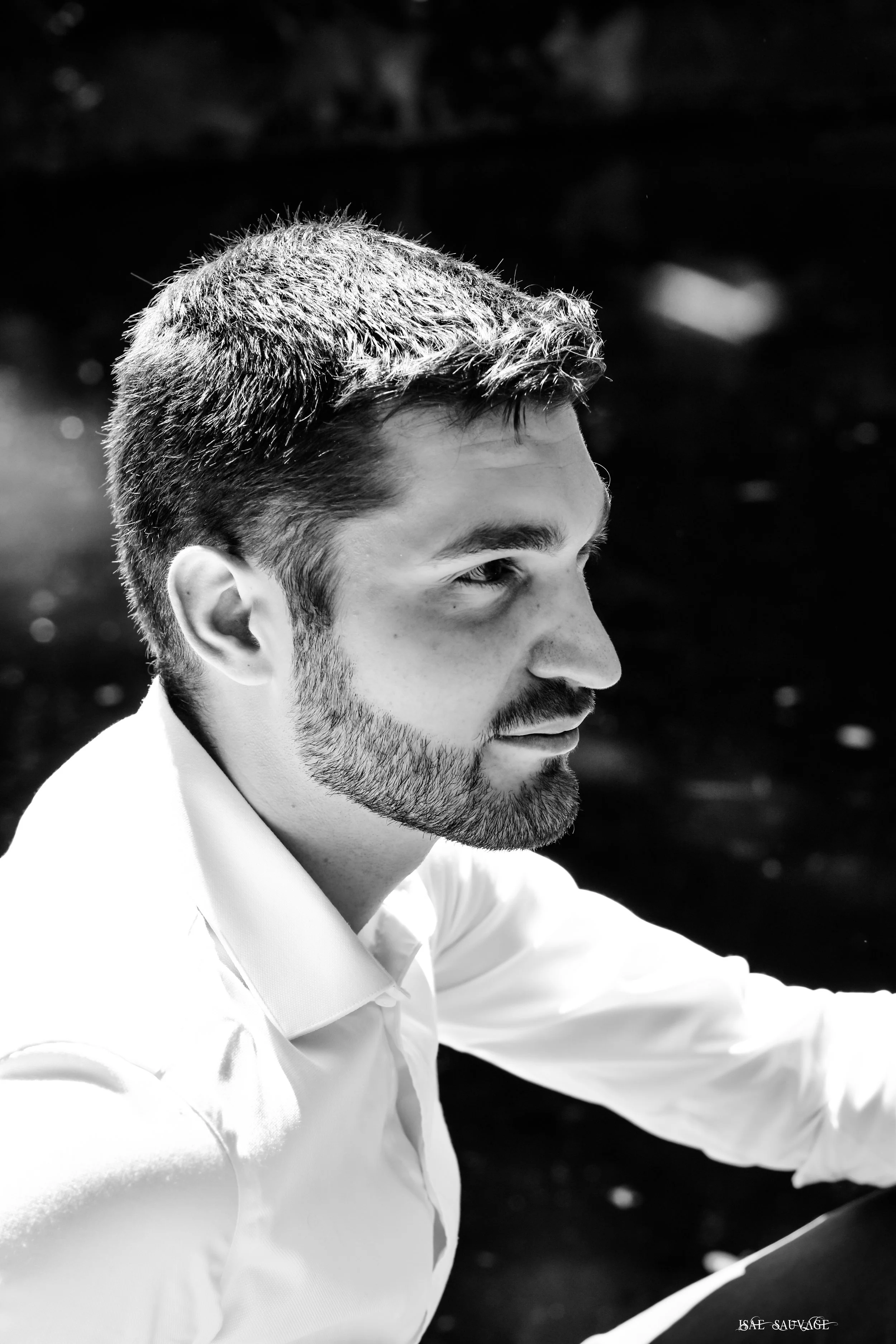 Portrait en noir et blanc d'un jeune homme avec des cheveux courts, une barbe légère, portant une chemise blanche, regardant vers la droite, avec un fond flou.