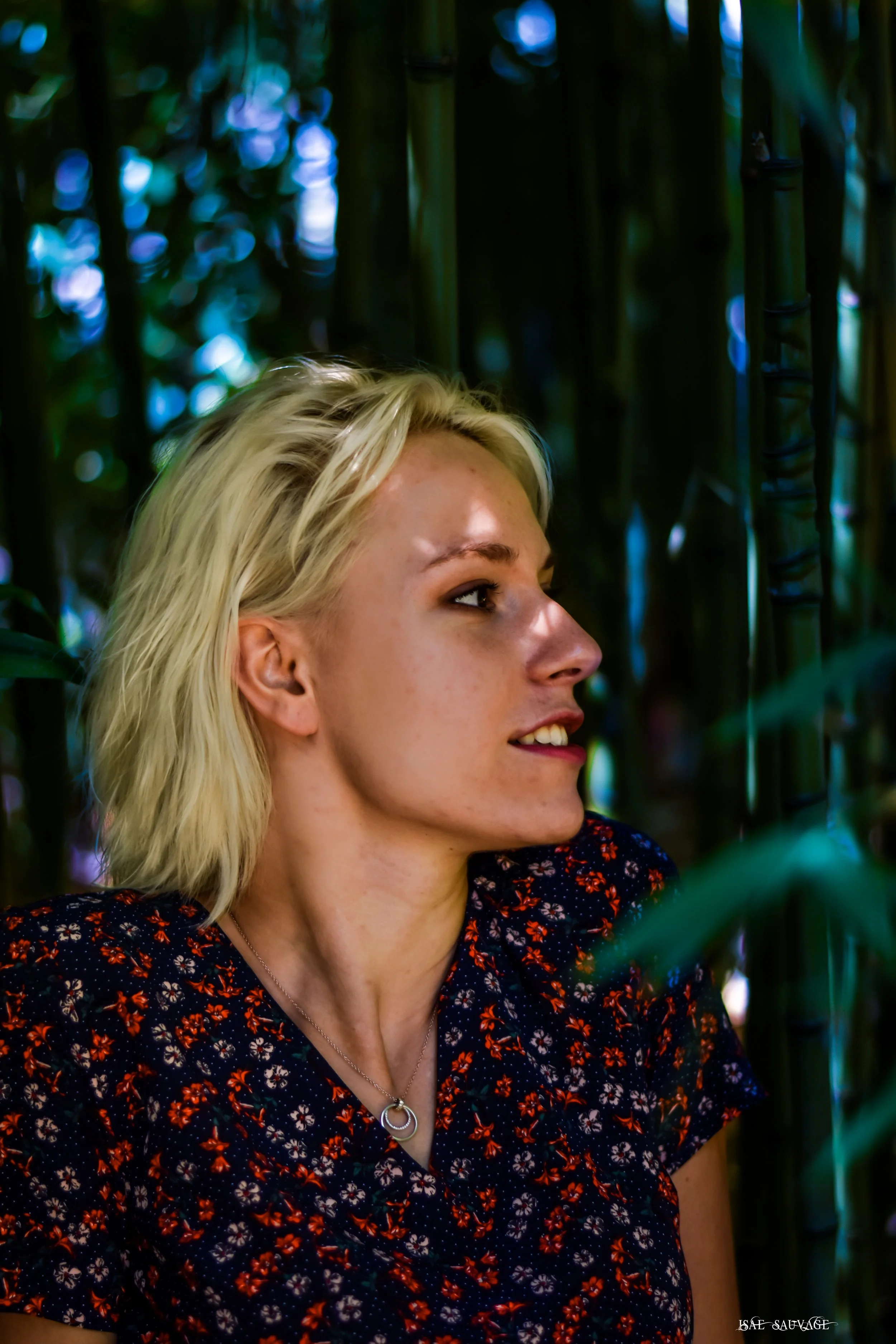 Portrait d'une femme blonde souriante portant un vêtement à motifs floraux rouge et noir, dans un environnement avec des tiges de bambou vertes en arrière-plan.