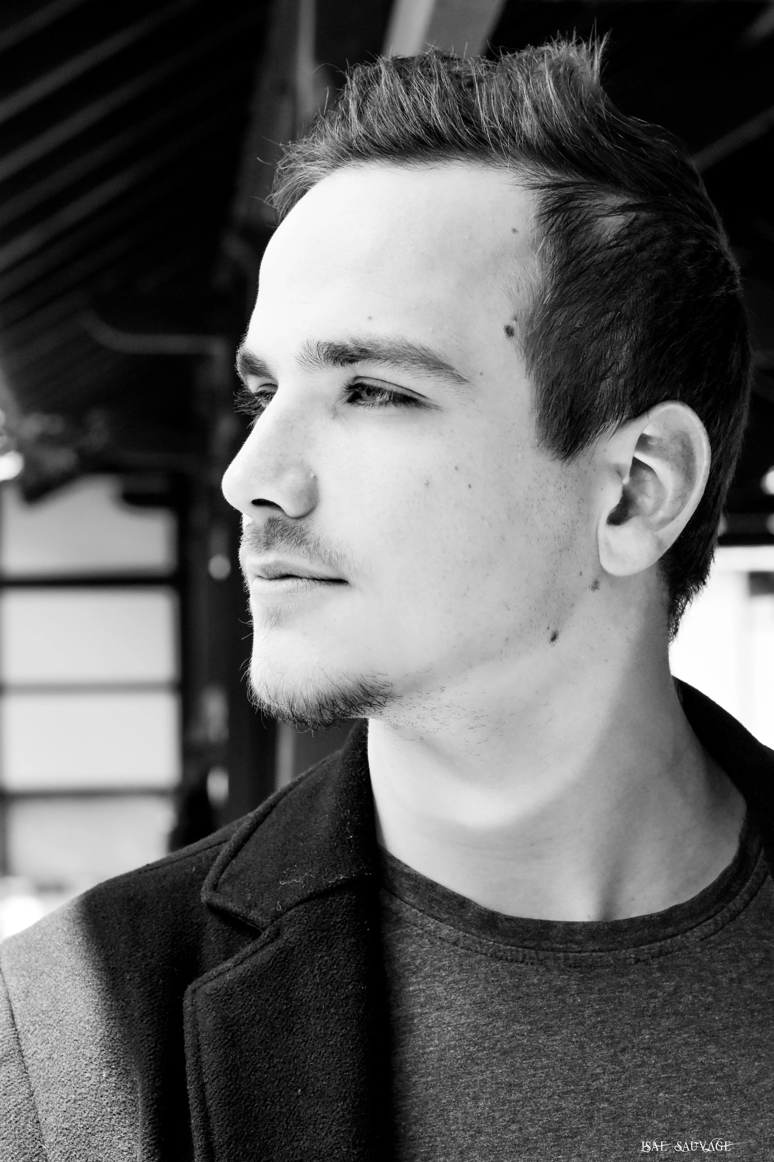 Portrait d'un jeune homme avec cheveux courts coiffés en arrière, regardant vers la gauche, portant un sweat à capuche noir, dans un environnement extérieur. 