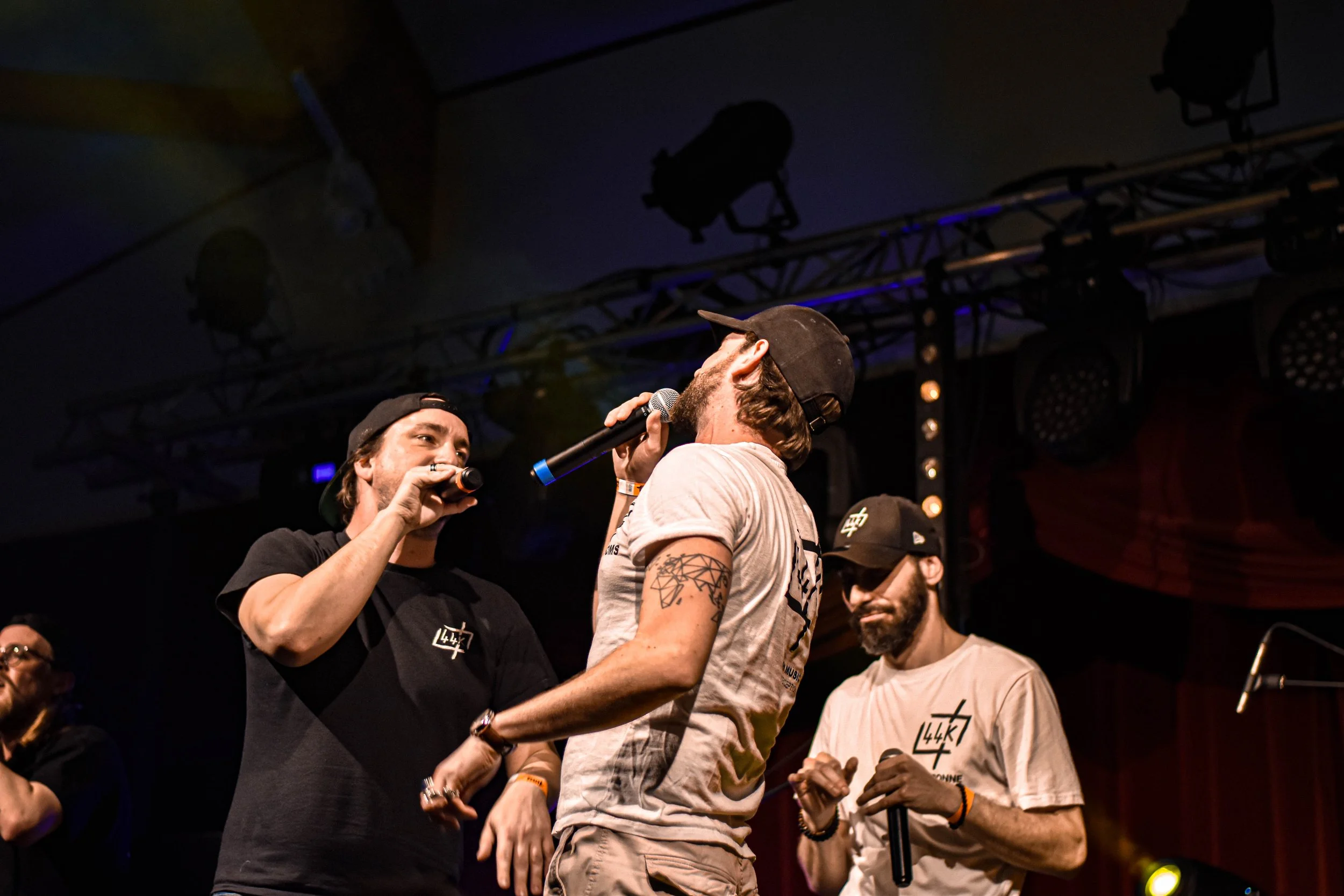 Deux hommes sur scène, l'un tenant un microphone, chantent devant un public dans une ambiance de concert. Il y a des équipements d’éclairage suspendus au plafond et d'autres personnes en arrière-plan.