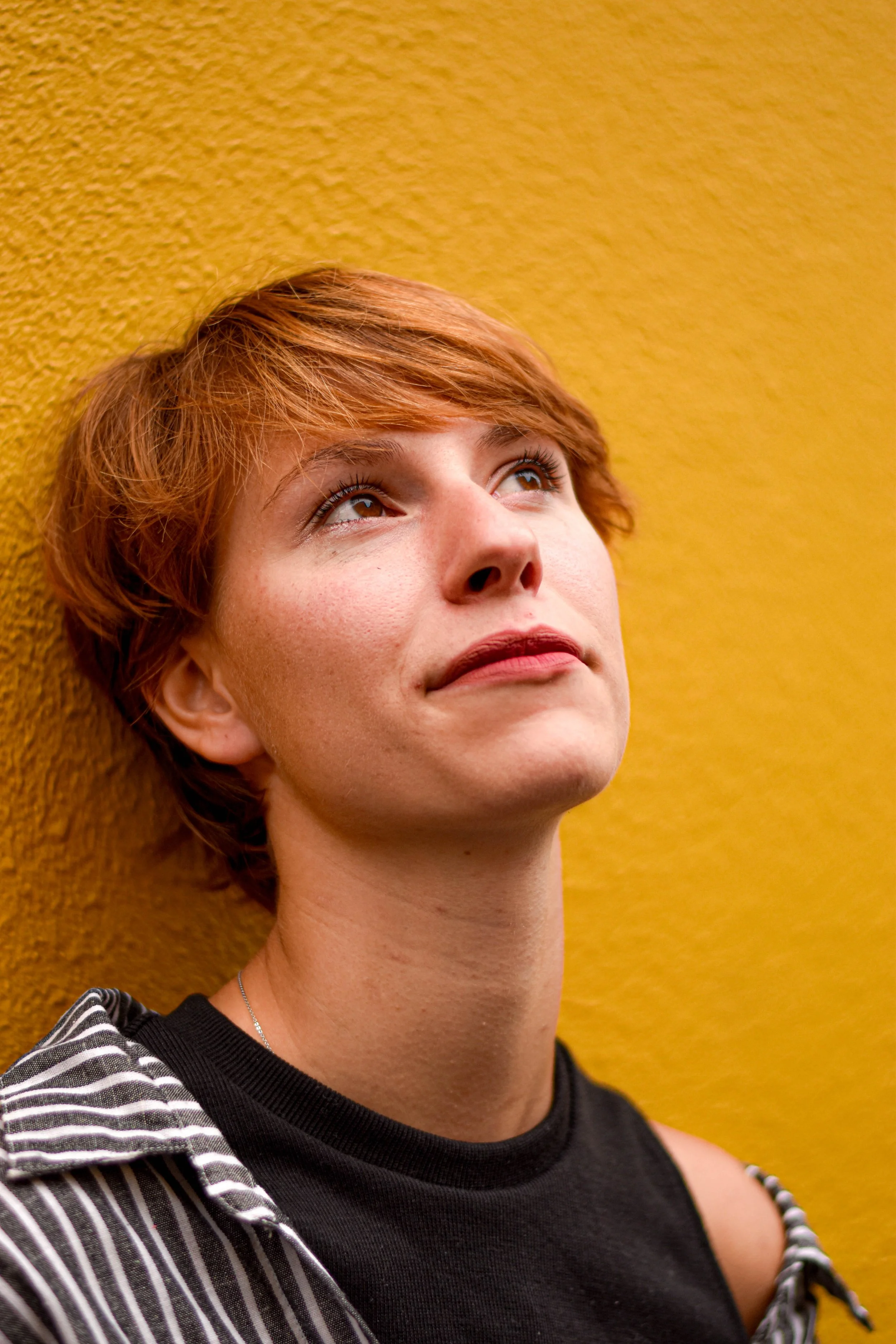 Une femme aux cheveux courts et roux, portant un t-shirt noir et une veste à rayures, regarde vers le haut avec un fond jaune.