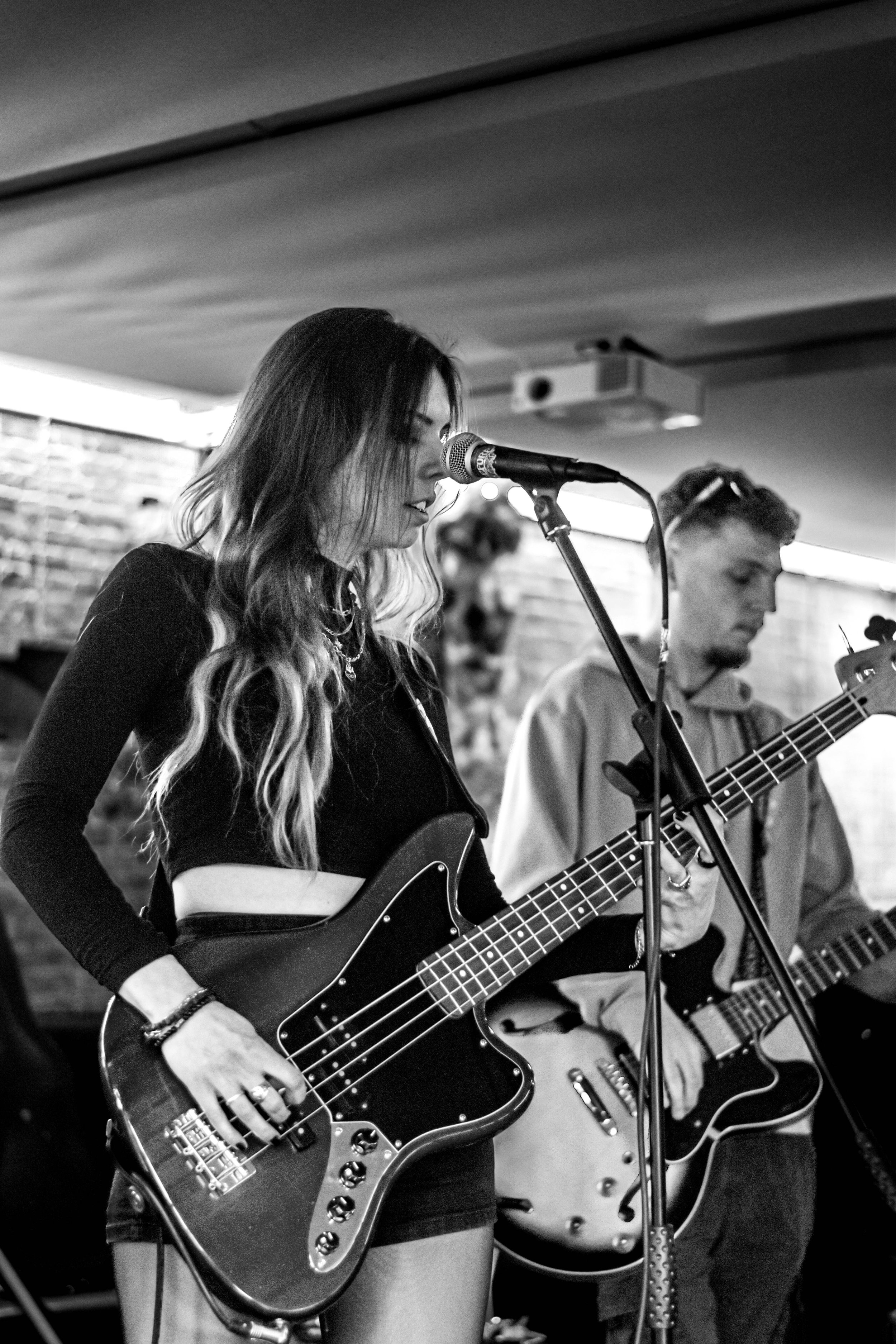 Une jeune femme joue de la guitare électrique en chantant dans bar, accompagnée d'un homme également en train de jouer de la guitare électrique, avec un micro devant elle.