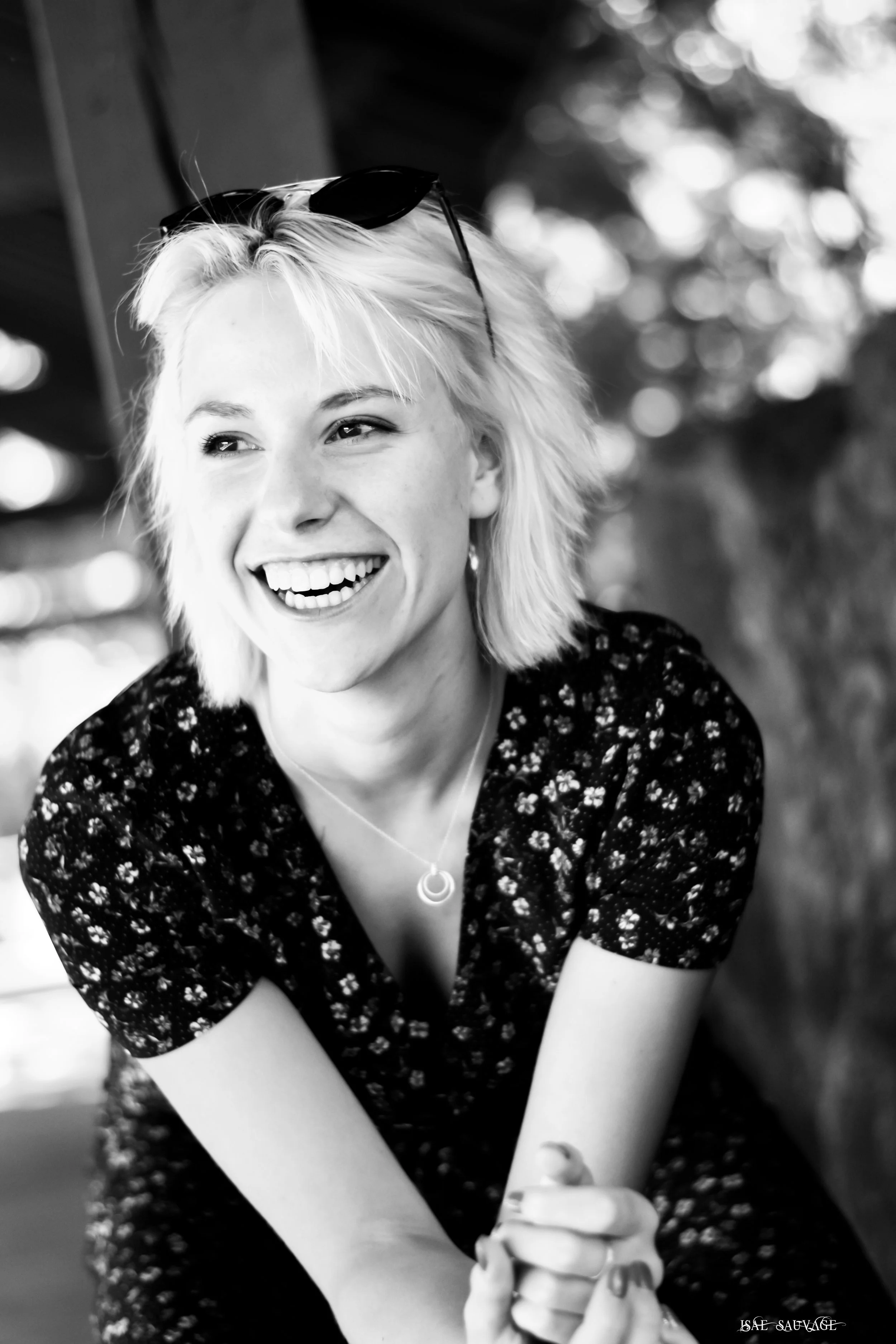 Jeune femme aux cheveux blonds courts, souriante, portant des lunettes sur la tête, vêtue d'une robe à motifs floraux, avec un collier, posant sourire à l'extérieur.