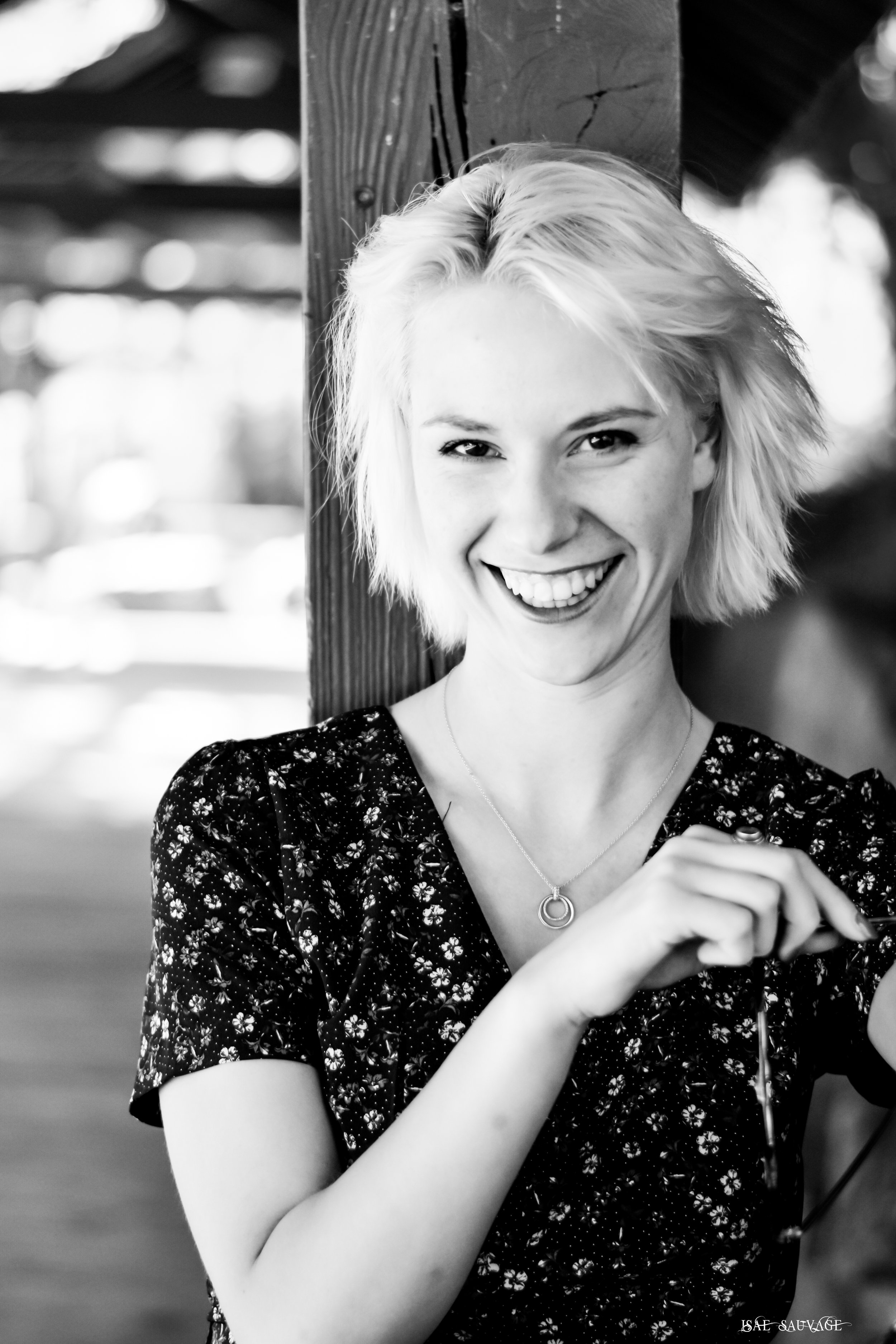 Une jeune femme souriante à cheveux blonds courts, portant un vêtement à motif floral, pose devant une structure en bois, probablement à l'extérieur.