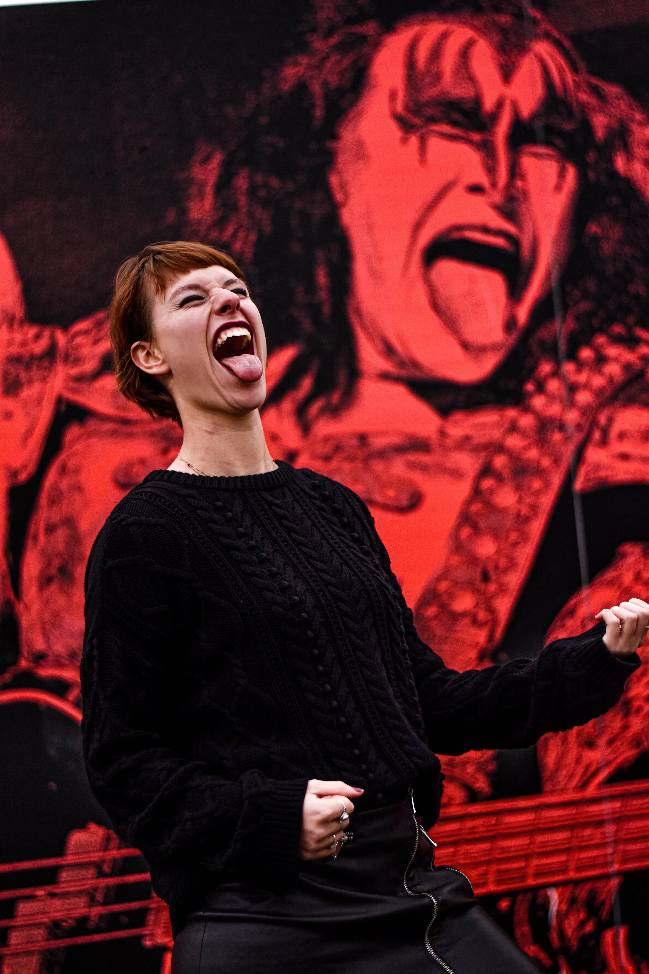 Une femme avec les cheveux courts roux, portant un pull noir, imitant un personne en train de jouer de la guitare électrique devant une affiche du groupe Kiss.