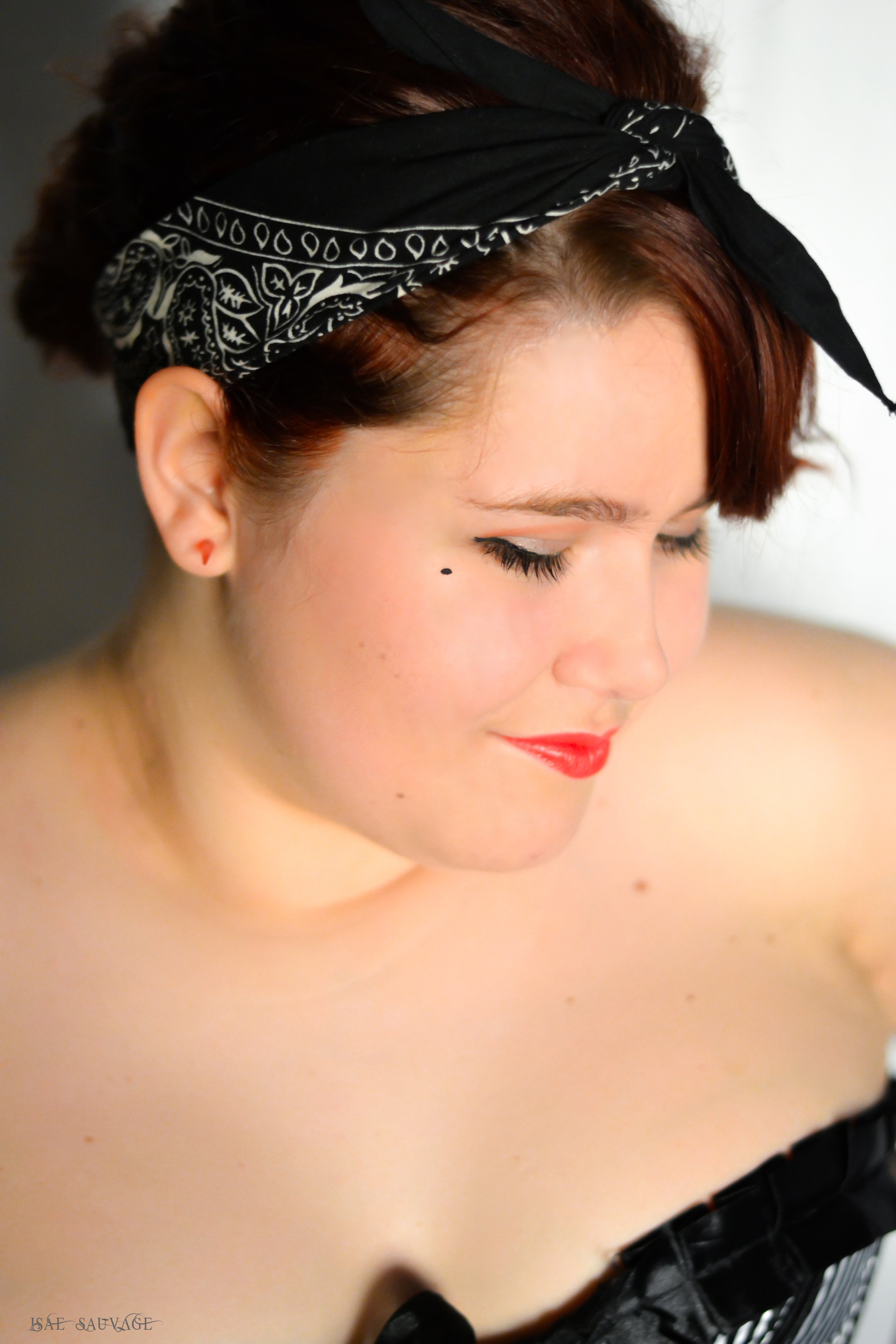 Une femme avec des cheveux relevés roux, portant un bandana noir avec un motif blanc, regarde vers le bas, avec un maquillage rouge à lèvres et un point noir près de l'œil, sans vêtements visibles. Portrait en gros plan.