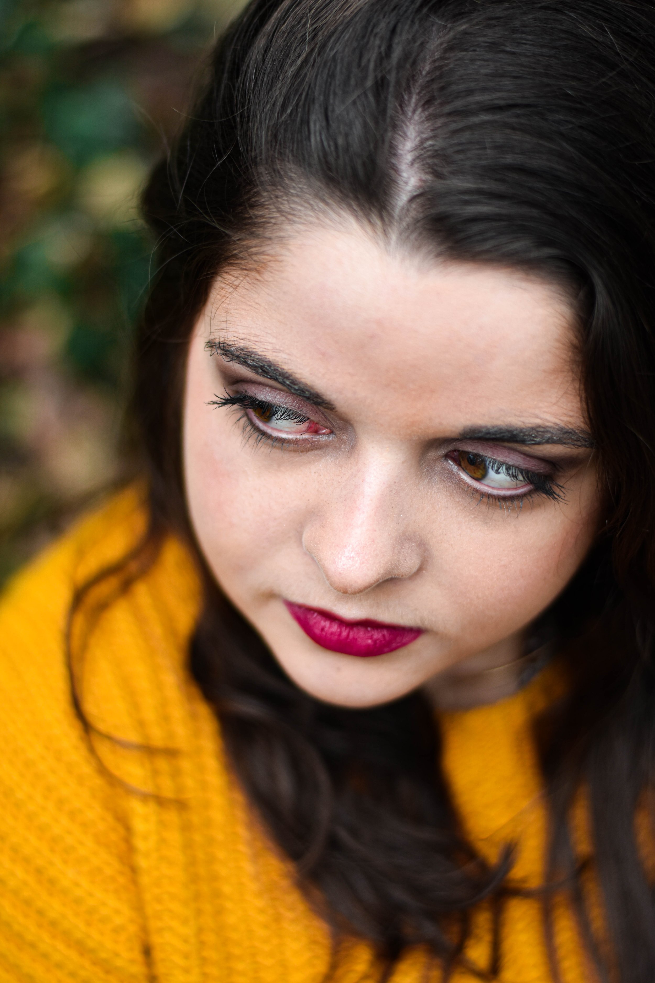 Un gros plan du visage d'une femme avec des cheveux bruns, portant un maquillage avec des lèvres roses et un pull jaune, dans un cadre naturel extérieur.