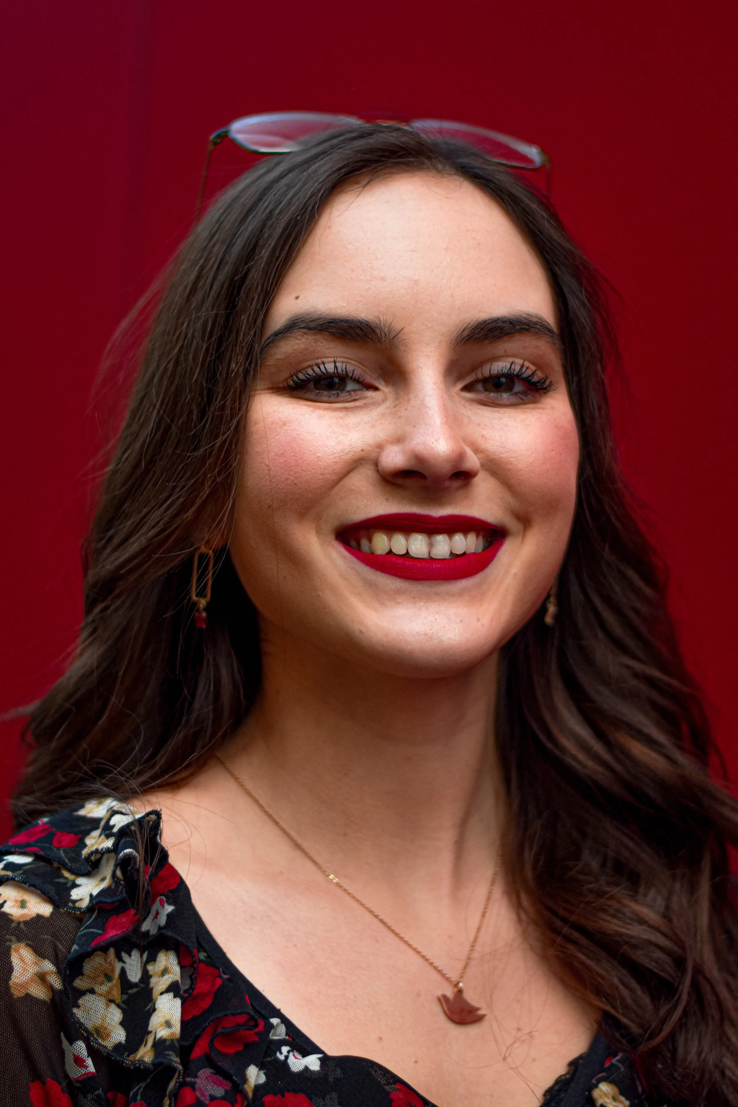 Jeune femme souriante avec des cheveux longs bruns, portant des lunettes sur la tête, un collier et un maquillage rouge, portant un haut à motifs floraux, près d'un fond rouge.