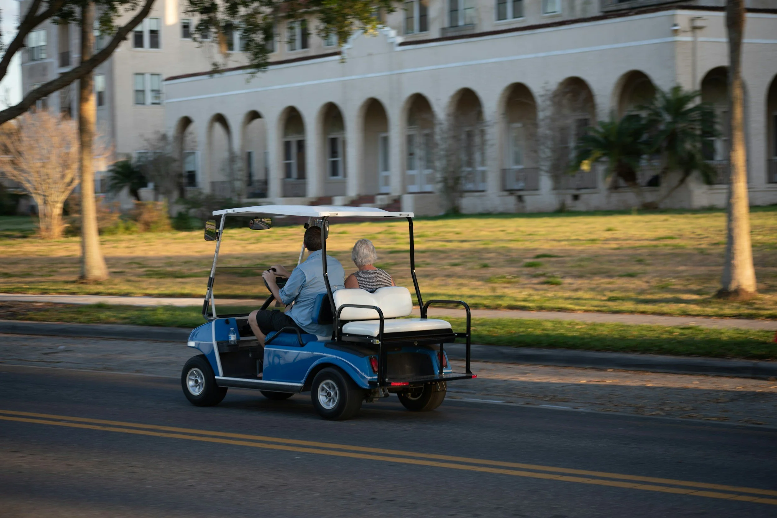 A man and an older woman riding in a blue golf cart on a street in a residential area with large white apartment buildings and trees in the background.