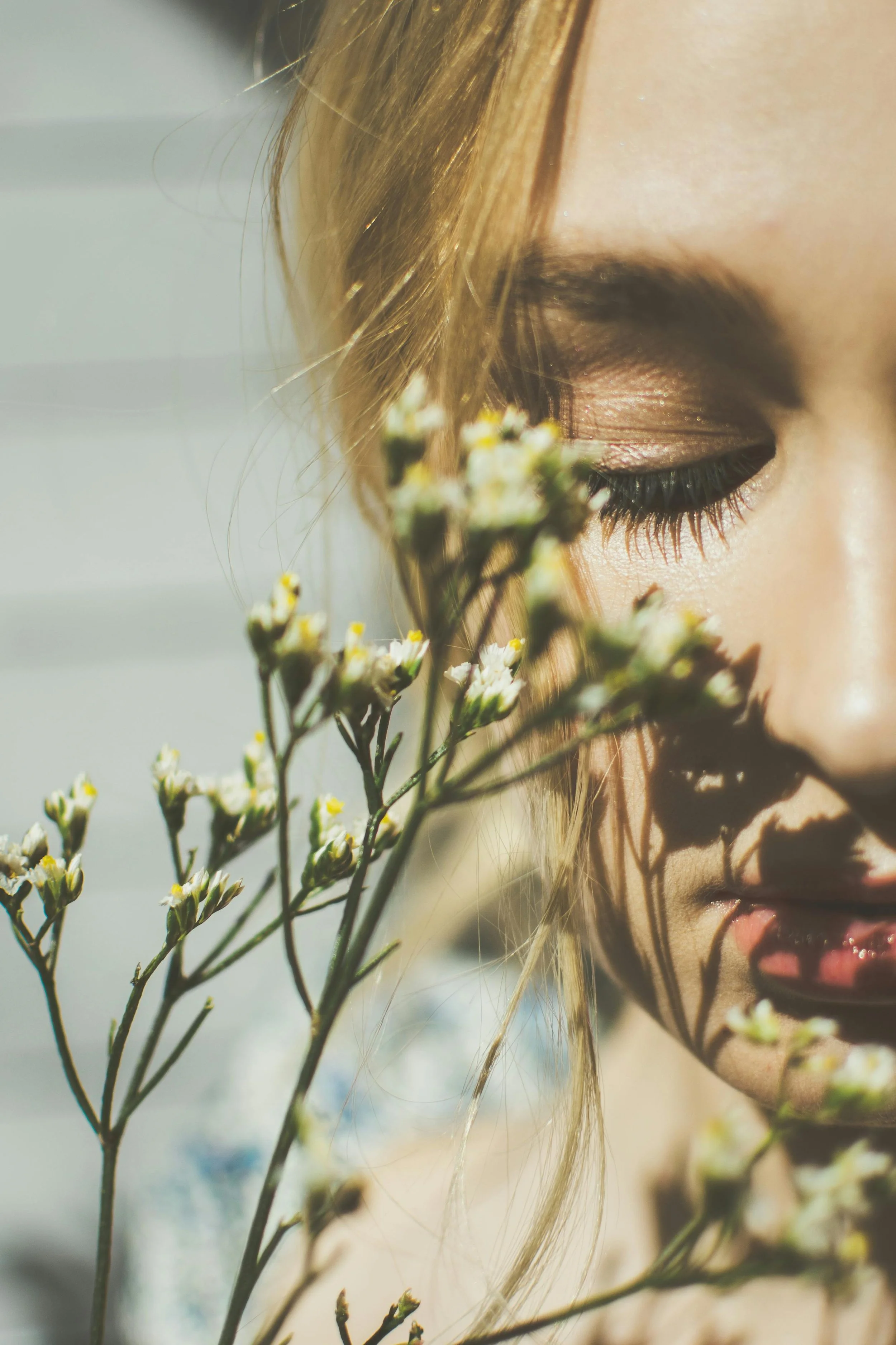 Close-up of a woman's face with eyes closed, holding small white and yellow flowers near her face with shadows cast from the flowers.
