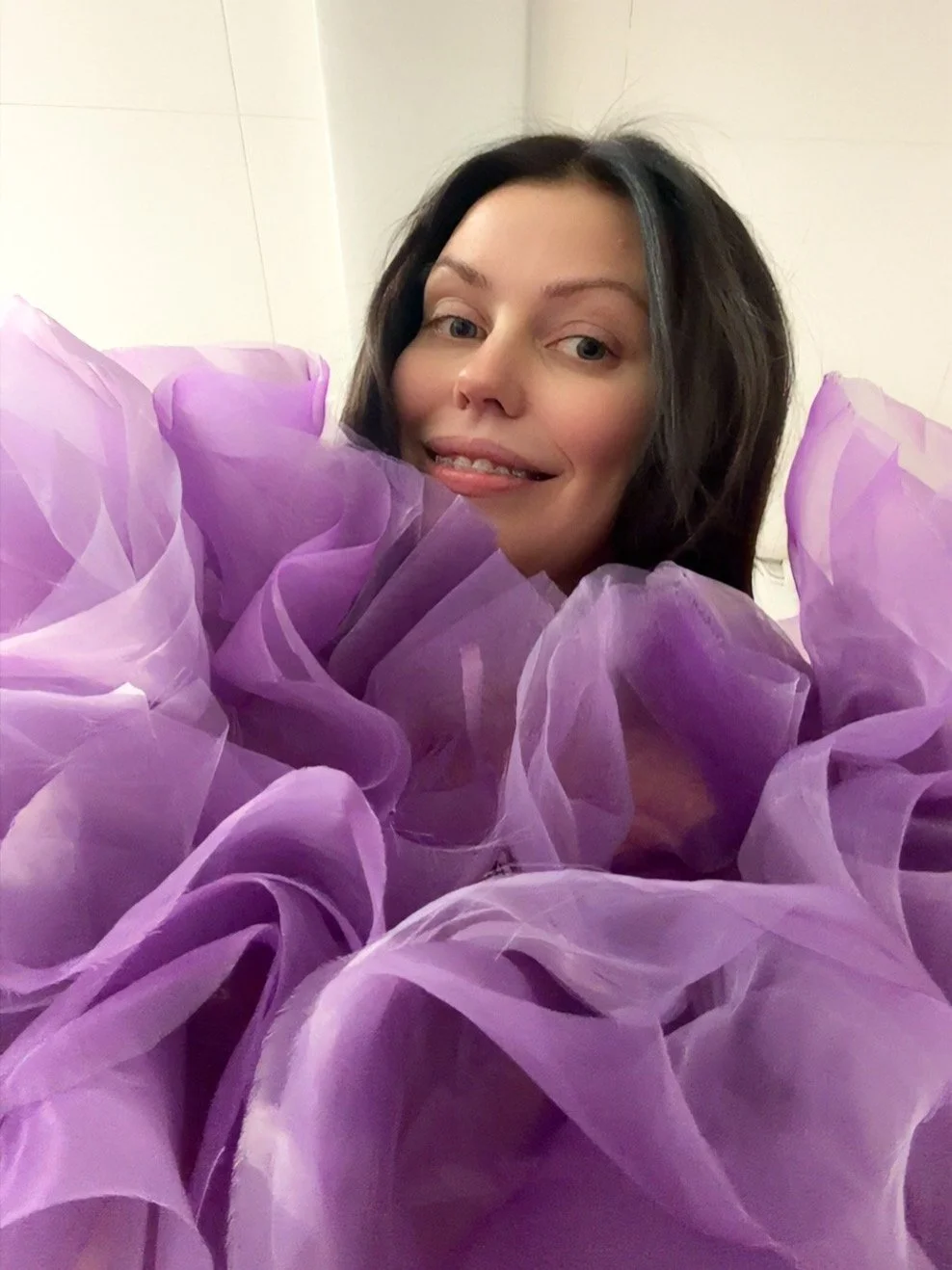A woman smiling behind large purple ruffled fabric or paper decorations.