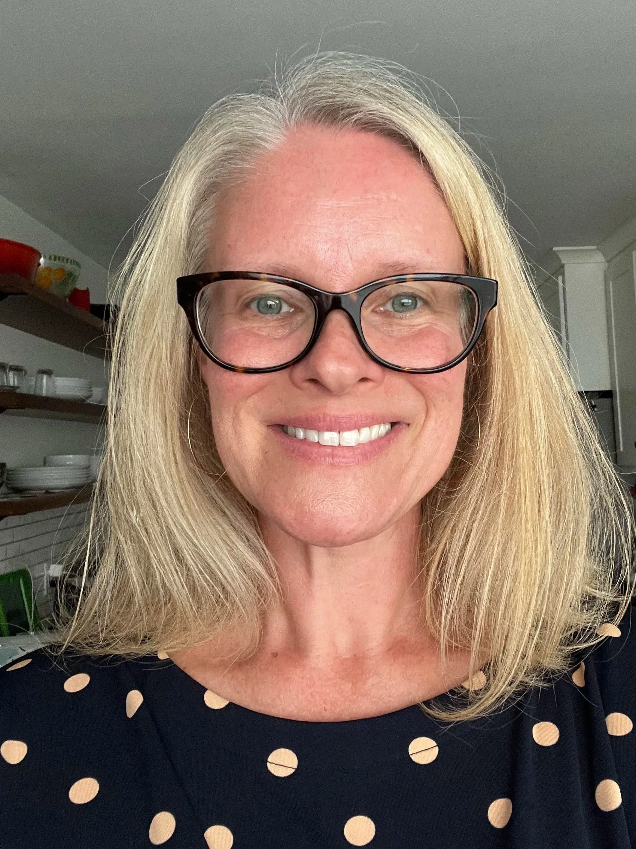 A woman with blonde hair, glasses, and a black dress with tan polka dots smiling indoors in a kitchen.