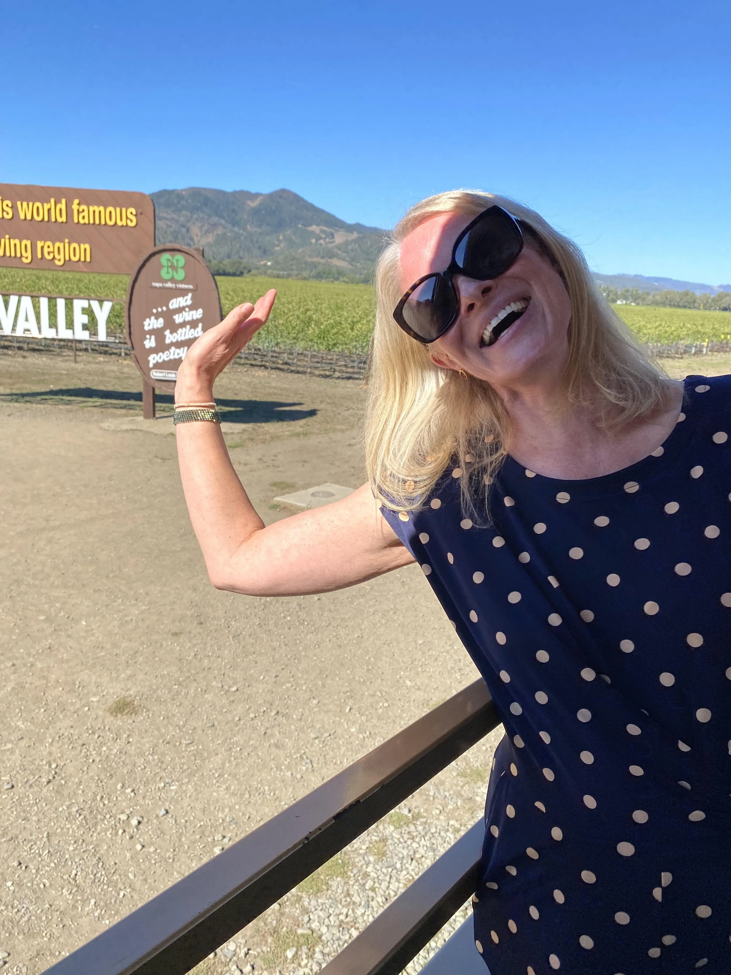 A smiling woman wearing sunglasses and a navy polka dot shirt pointing to a sign in a vineyard with mountains in the background.
