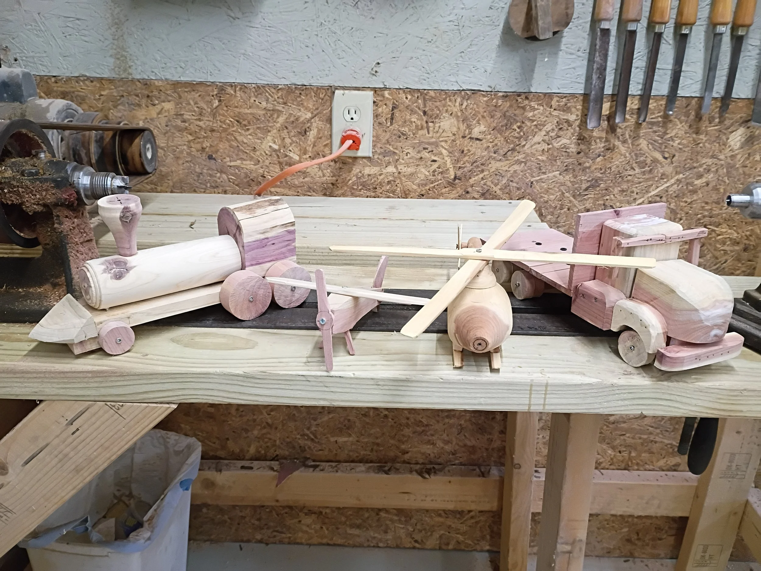 Wooden models of various vehicles including a train, helicopter, and truck on a workbench in a workshop.