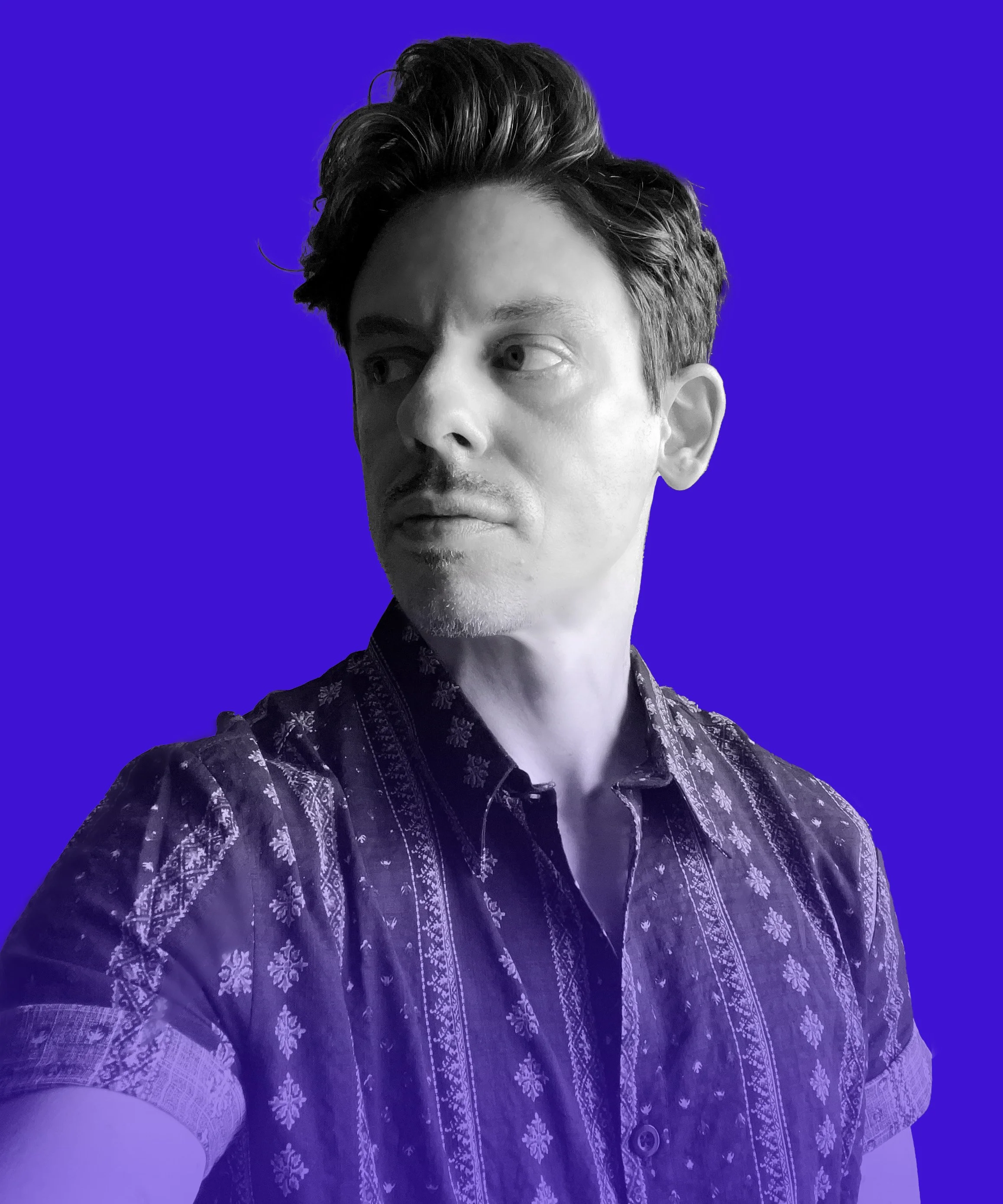 A black and white photo of a man with dark, styled hair, looking to the side, against a blue background, wearing a patterned shirt.