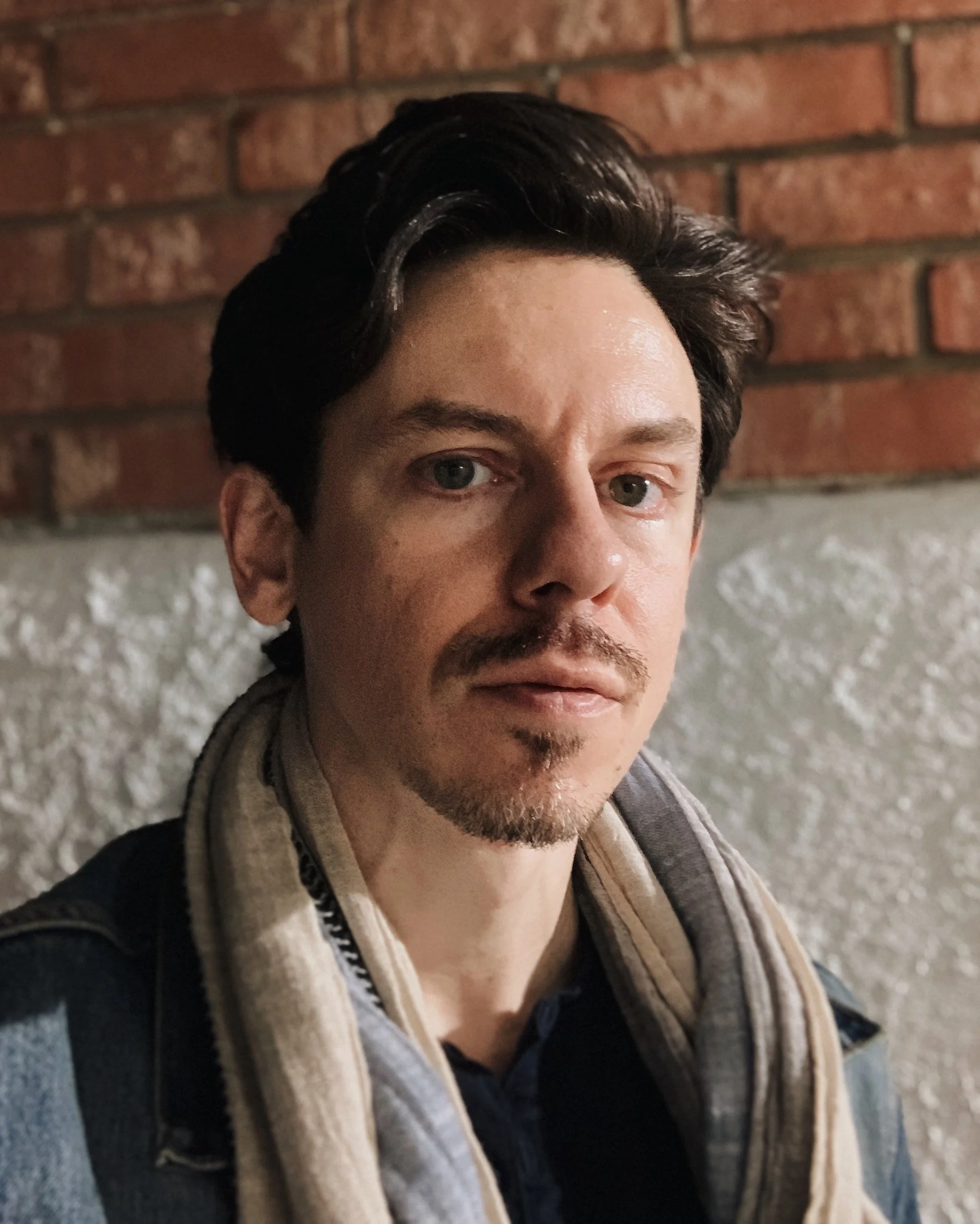 A young man with dark hair and a small beard, wearing a denim jacket, a scarf, and a backpack, standing in front of a brick and concrete wall.