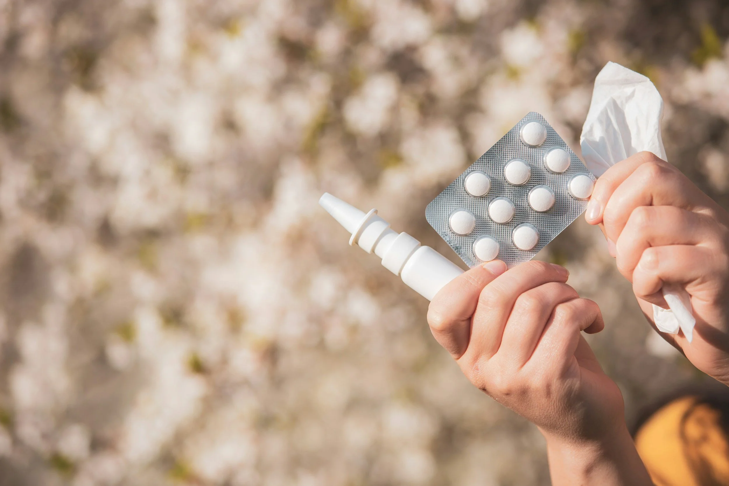 Hände halten eine Packung Tabletten, eine Nasenspray-Flasche und ein zerknülltes Papiertuch. Hintergrund unscharf mit blühenden Bäumen.