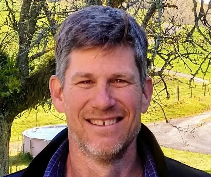 A smiling middle-aged man with short, graying hair and a beard, outdoors in a rural setting with a tree and grassy landscape in the background.