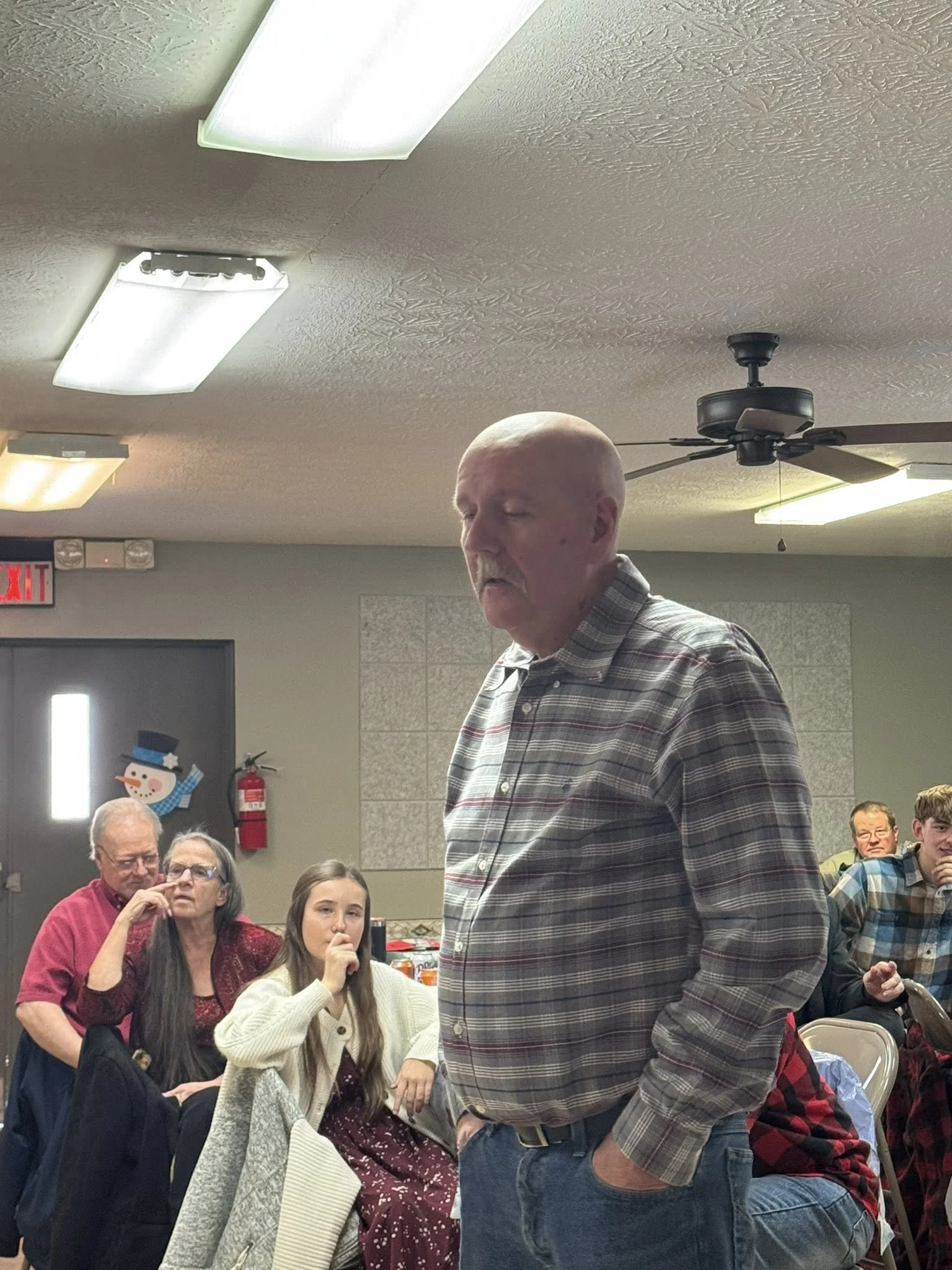 A man with a bald head and mustache in a plaid shirt stands in front of a seated audience in a room with a Christmas decoration and a snowman on the door.