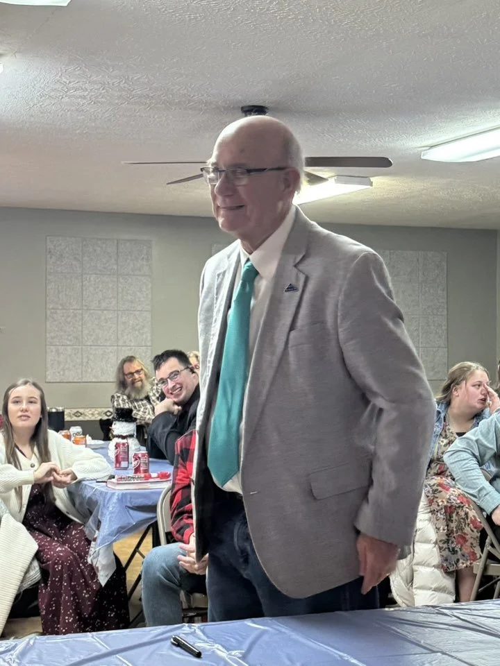 An older man with glasses, bald head, and a gray suit with a turquoise tie standing in a room with other seated people at tables, celebrating or attending an event.