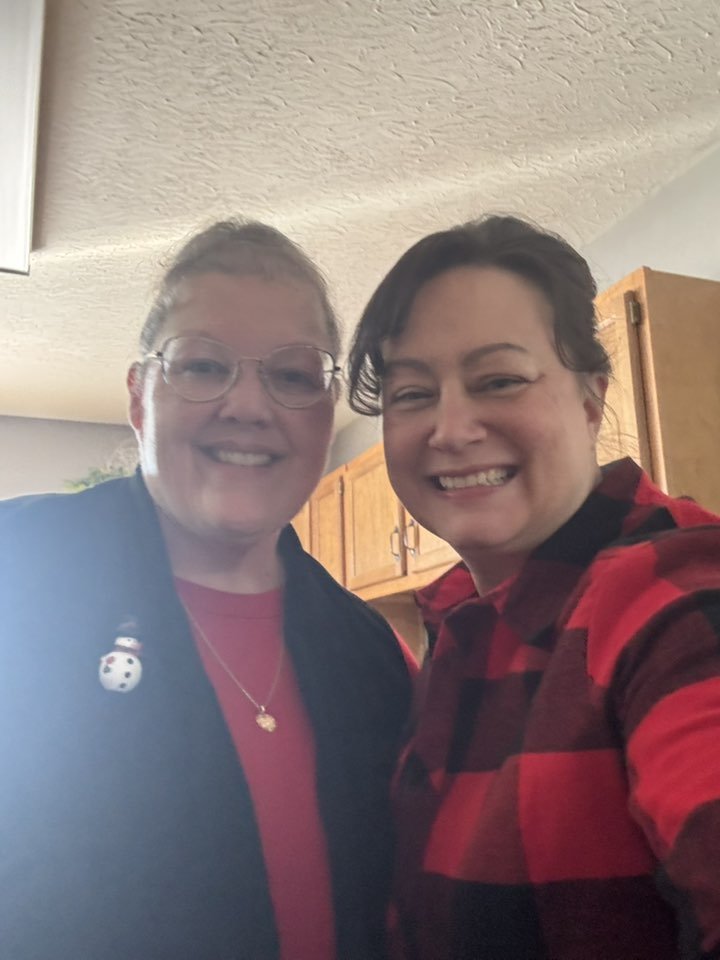 Two women smiling for a selfie in a kitchen, one wearing glasses and a black jacket with a snowman pin, the other wearing a red and black plaid shirt.