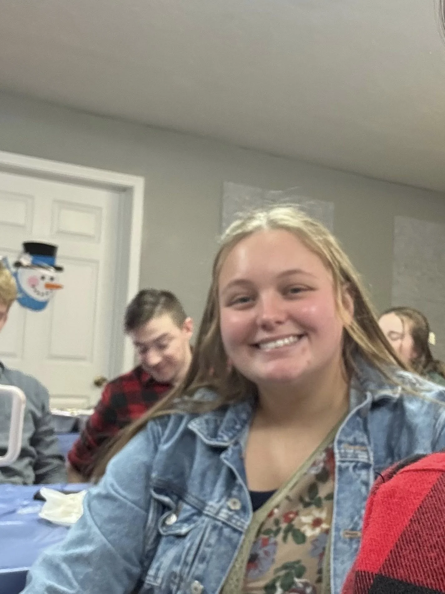 A group of young people at a holiday gathering, with a smiling girl in the foreground and a snowman decoration on the wall in the background.