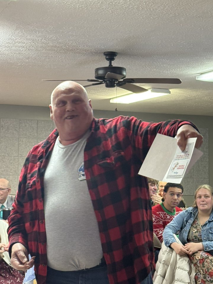 A man in a red and black plaid shirt holding a piece of paper, smiling, in a room with other people seated and a ceiling fan overhead.