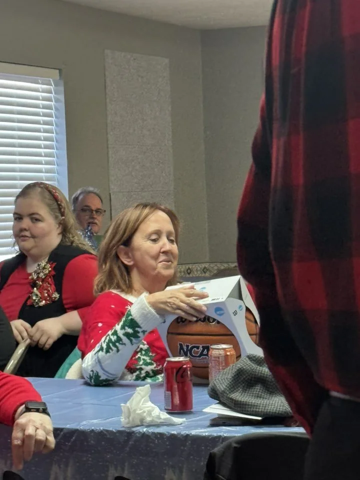 A woman in a festive Christmas sweater opening a gift at a holiday gathering, with other people sitting nearby.