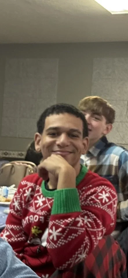 A young man in a red holiday sweater with white snowflakes and green cuffs, smiling and resting his chin on his hand, at a gathering or party with other people in the background.