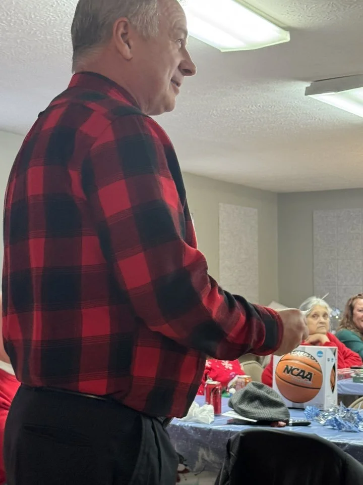 A man in a red and black plaid shirt standing at a gathering with women seated at a table, with a basketball-themed gift box and cans of soda visible on the table.