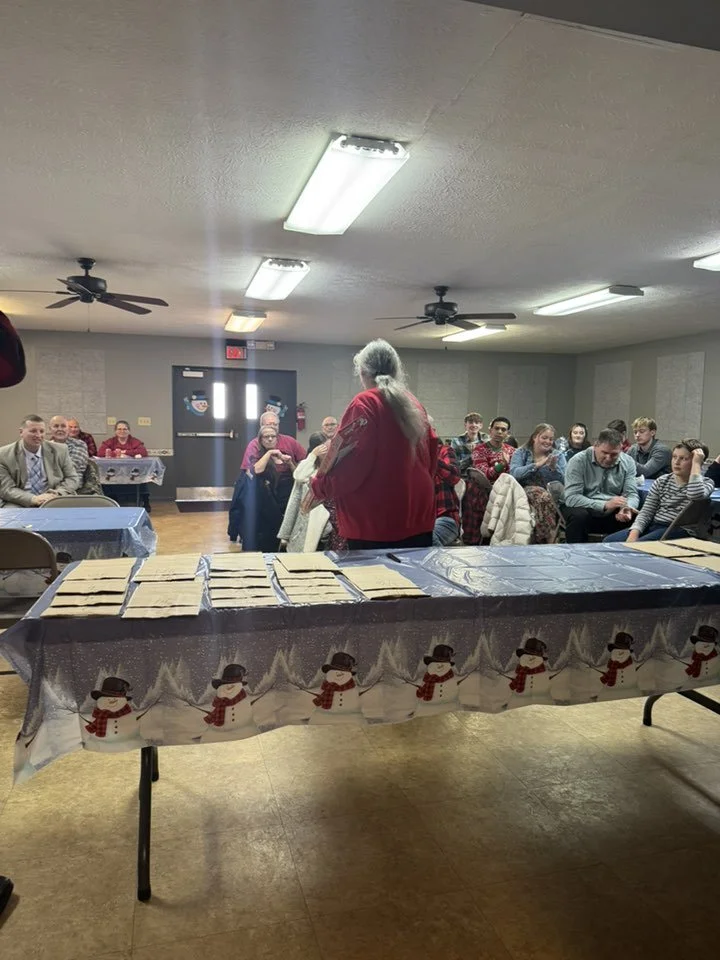 A group of people seated at tables in a room with Christmas decorations, facing a woman standing at the front. The front table is decorated with a snowman-themed tablecloth and has several lined-up cookies on it.