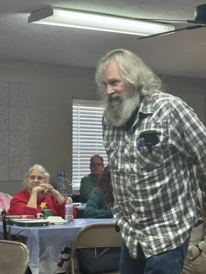 An elderly man with long gray hair and a gray beard stands smiling in a room with festive holiday decorations, with several seated people in the background.