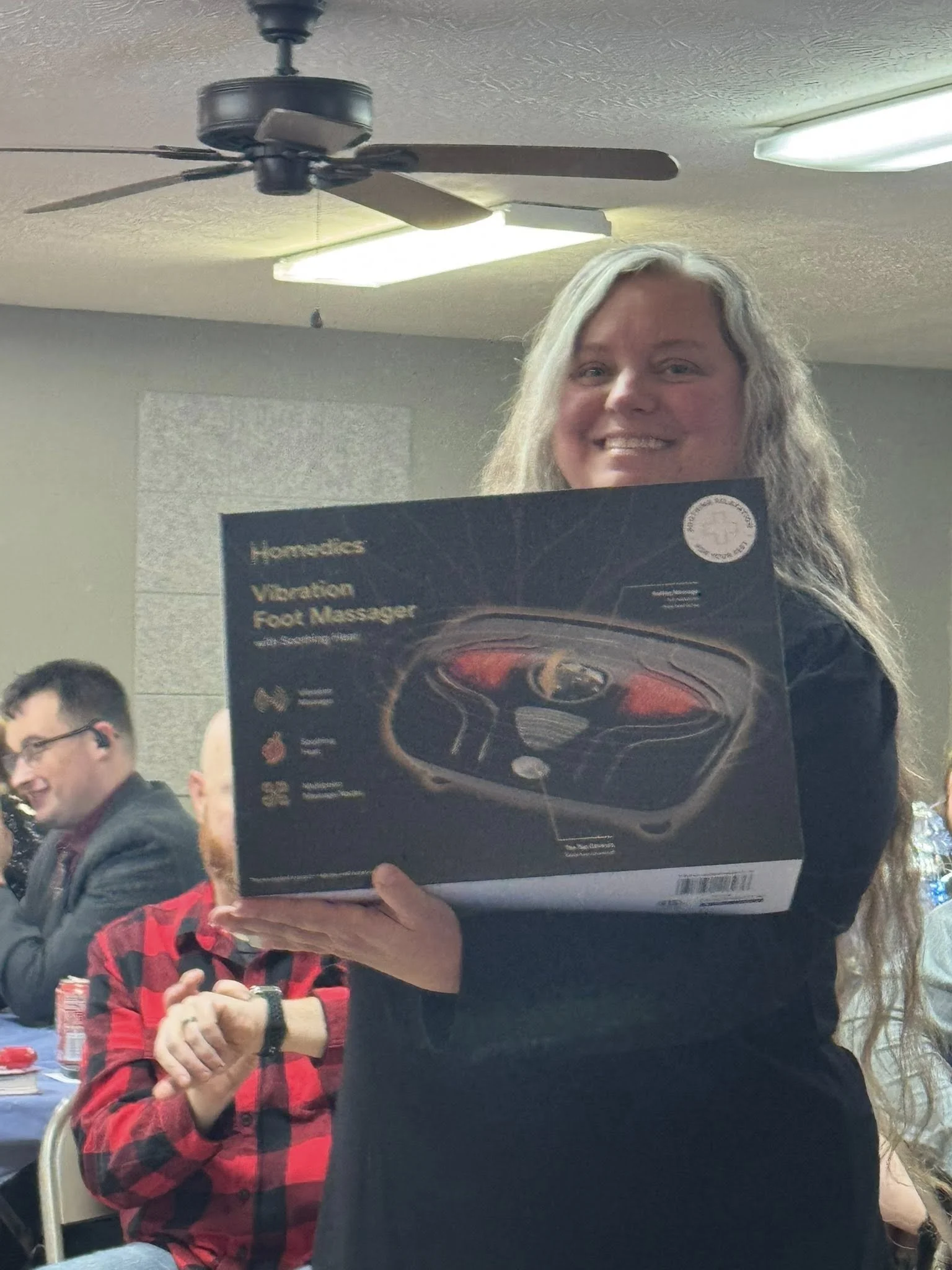 A woman with long gray hair smiling while holding a boxed home vibration foot massager at a gathering or event, with several people seated at tables behind her.