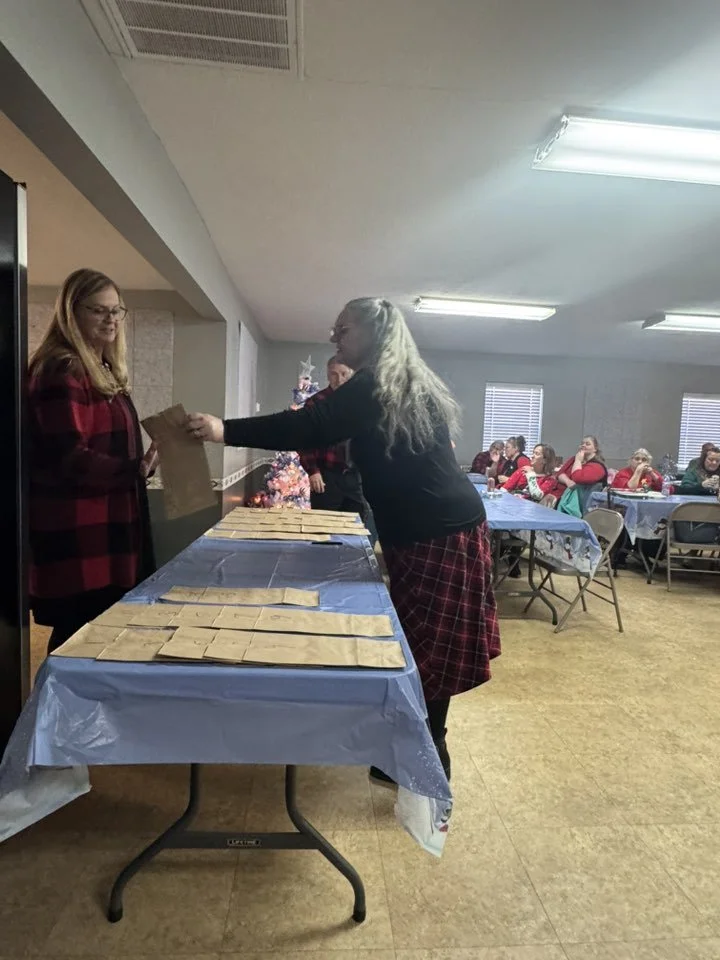 A woman in a black top and red plaid skirt receives a gift bag from another woman in a red and black checkered jacket at a Christmas gathering with a decorated tree and people sitting at tables in the background.