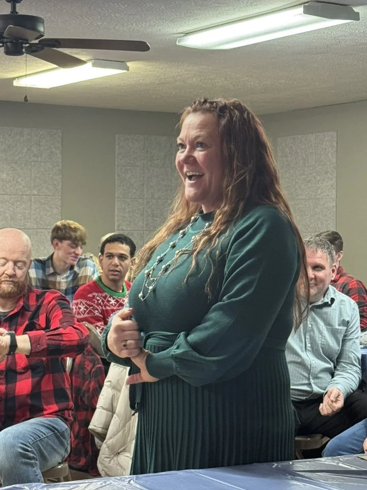 A woman with long wavy hair smiling and standing in front of a group of seated people in a room with grey walls and fluorescent lighting.