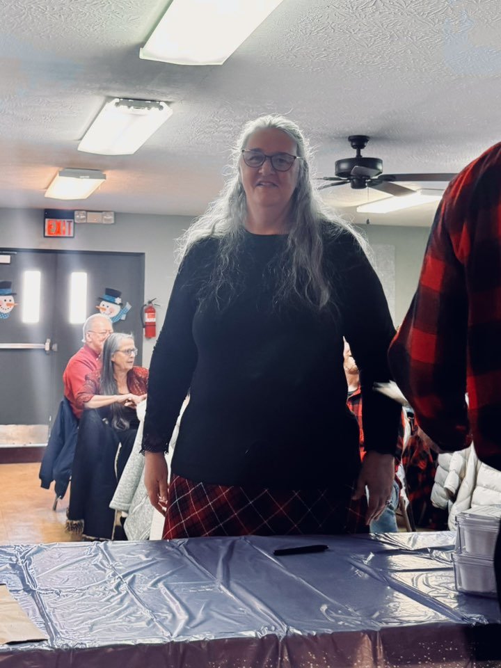 A woman with gray hair, glasses, and a black top standing at a table during a holiday gathering, with people sitting at tables and holiday decorations in the background.