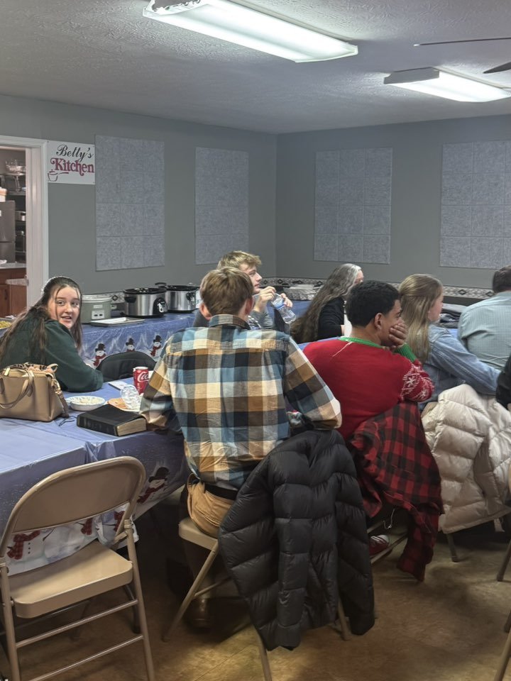 People sitting around a table at a gathering, with food and drinks, in a room decorated for a holiday or event.