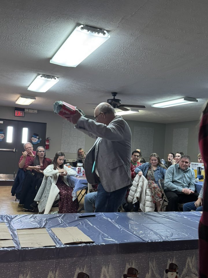 A man in a gray jacket is raising a wrapped gift above his head at a gathering with several seated people. The room has a ceiling fan, fluorescent lighting, and holiday snowman decorations on the door.