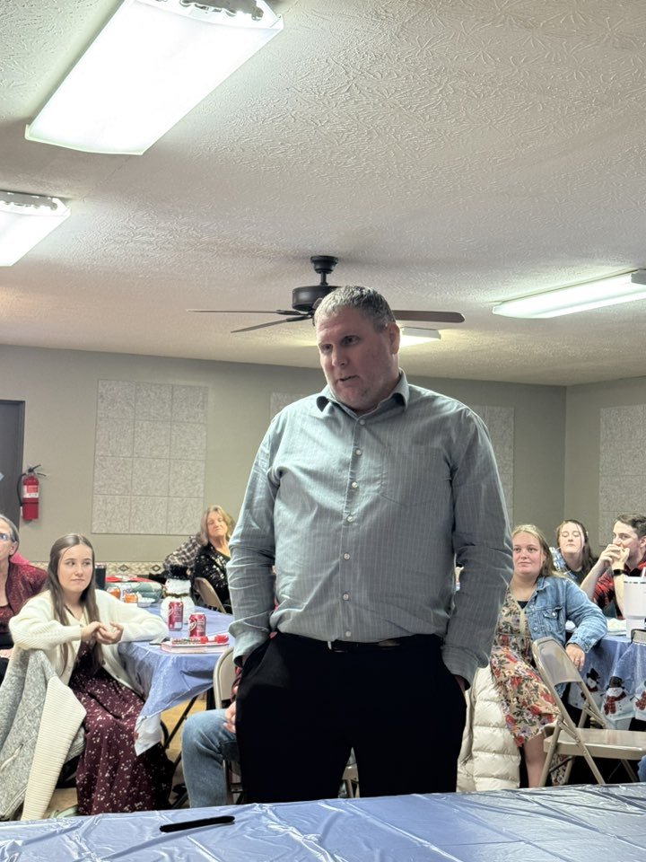 A man stands in front of a table with his hands in his pockets during a gathering or party, with several people seated at round tables decorated with holiday-themed tablecloths and drinks.