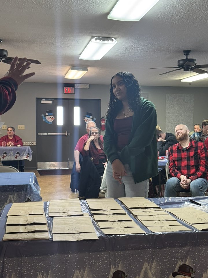 A woman with curly hair standing in front of a table with paper bags during a holiday gathering, with festive snowman decorations on the door in the background and several seated guests watching her.