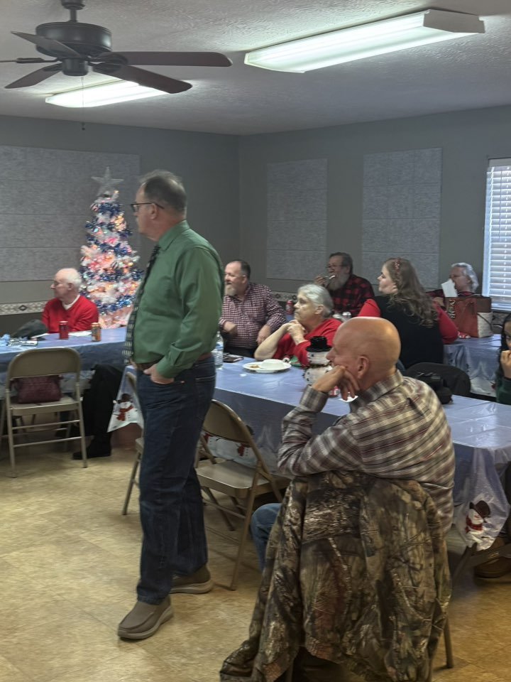 People gathered for a holiday celebration, seated at tables with holiday-themed tablecloths and decorations, including a decorated Christmas tree in the background.