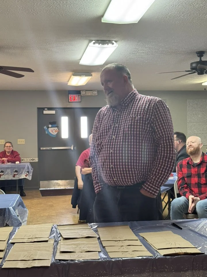 A group of people gathered in a room decorated for Christmas, with a man in a checkered shirt standing in front of a table covered with paper bags. The room has holiday decorations, including a snowman on the wall, and people seated at tables.