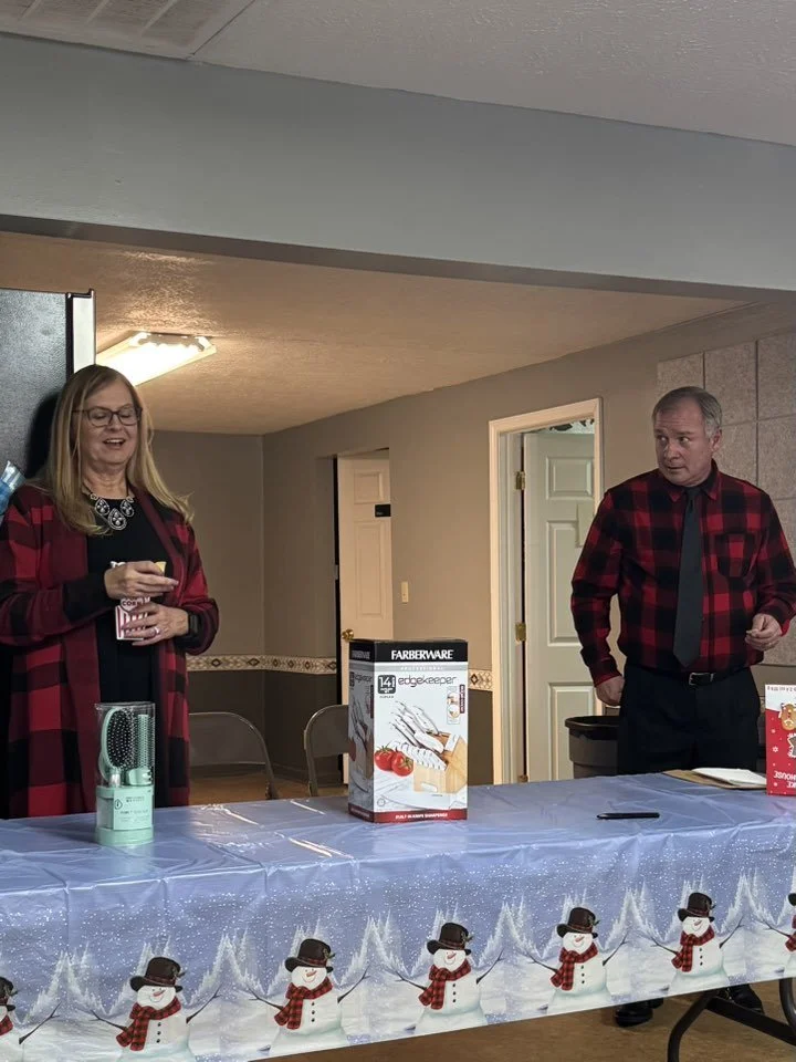 A woman and a man are standing behind a Christmas-themed decorated table, with snowmen illustrations on a blue tablecloth. The woman is holding a mug and the man is looking toward her. There are boxes and a portable fan on the table.