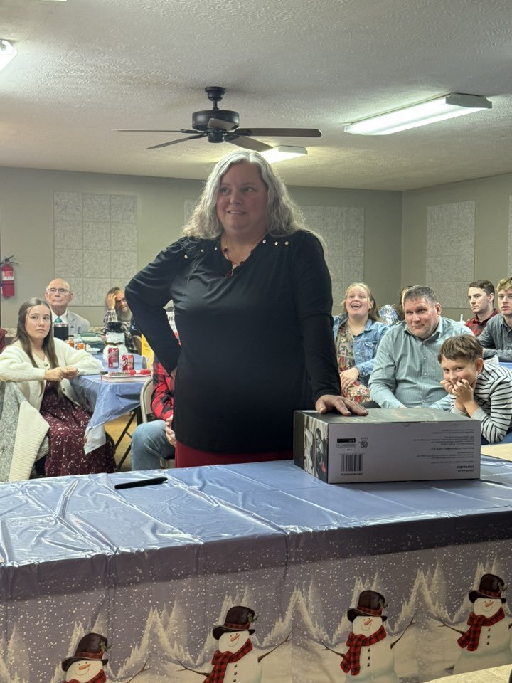 A woman in black standing at a table with a surprised expression, at a gathering with people sitting around. The table is decorated with a snowman-themed tablecloth, and there is a box and a black marker on it, in a room with a ceiling fan and fluore
