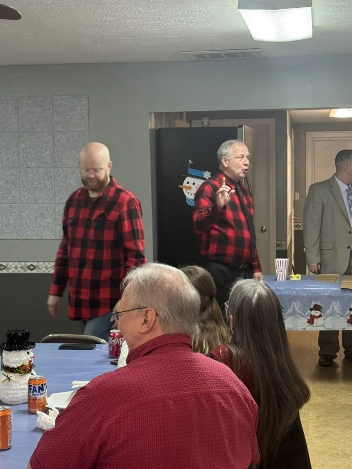 People gathered at a holiday party, with two men standing at the front wearing red and black plaid shirts, one speaking and the other smiling, while the attendees sit at tables decorated with snowmen and holiday decorations.